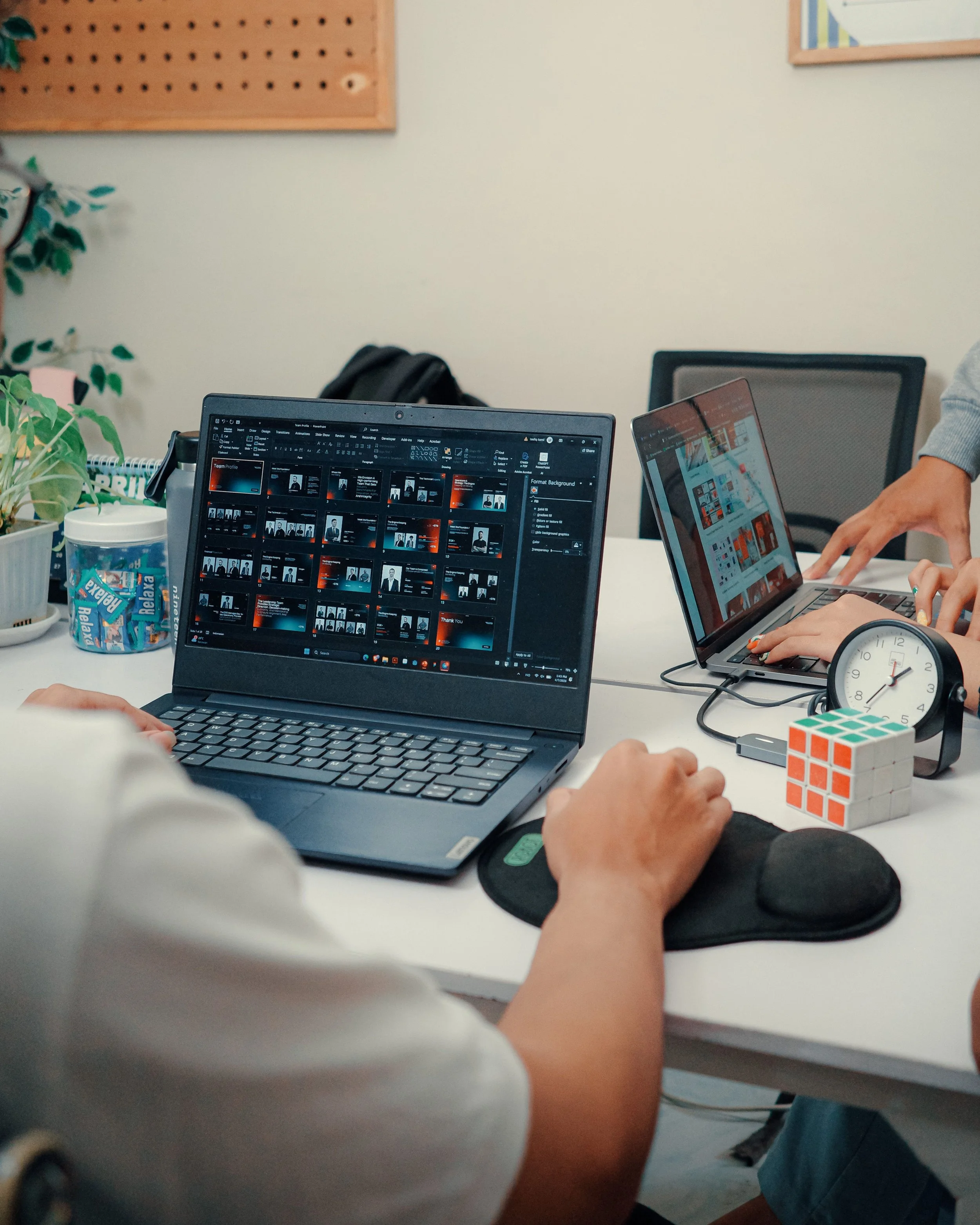 People working on laptops at a white table in an office, with a plant, a Rubik's cube, a clock, and a container of relief mixing tablets in the foreground.