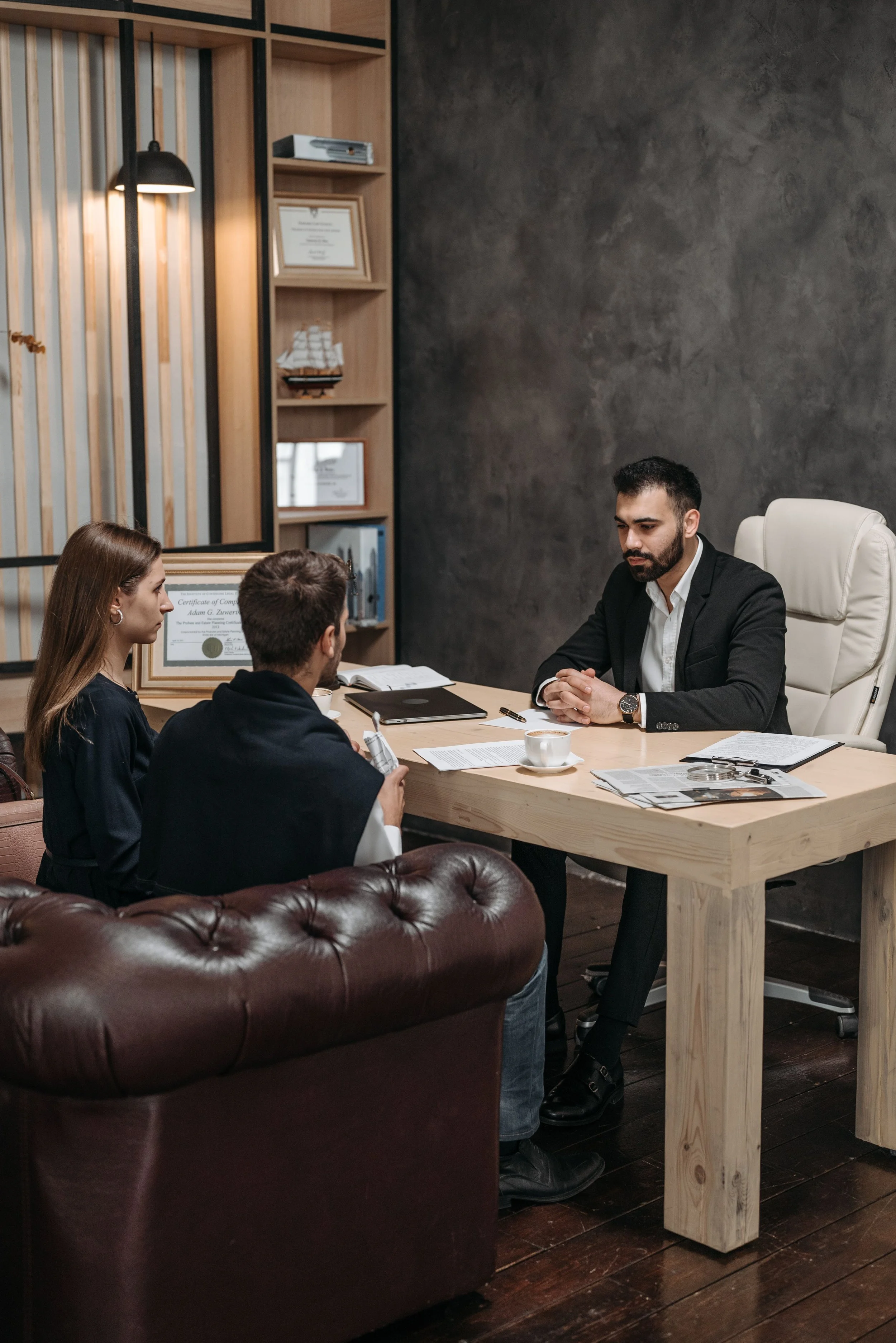 A business meeting taking place in a modern office with four people. The man in a suit is sitting at the head of the wooden table, with three others, including a woman and a man, facing him. The room has a dark textured wall, a leather couch, and wooden shelves with certificates, books, and a model ship.