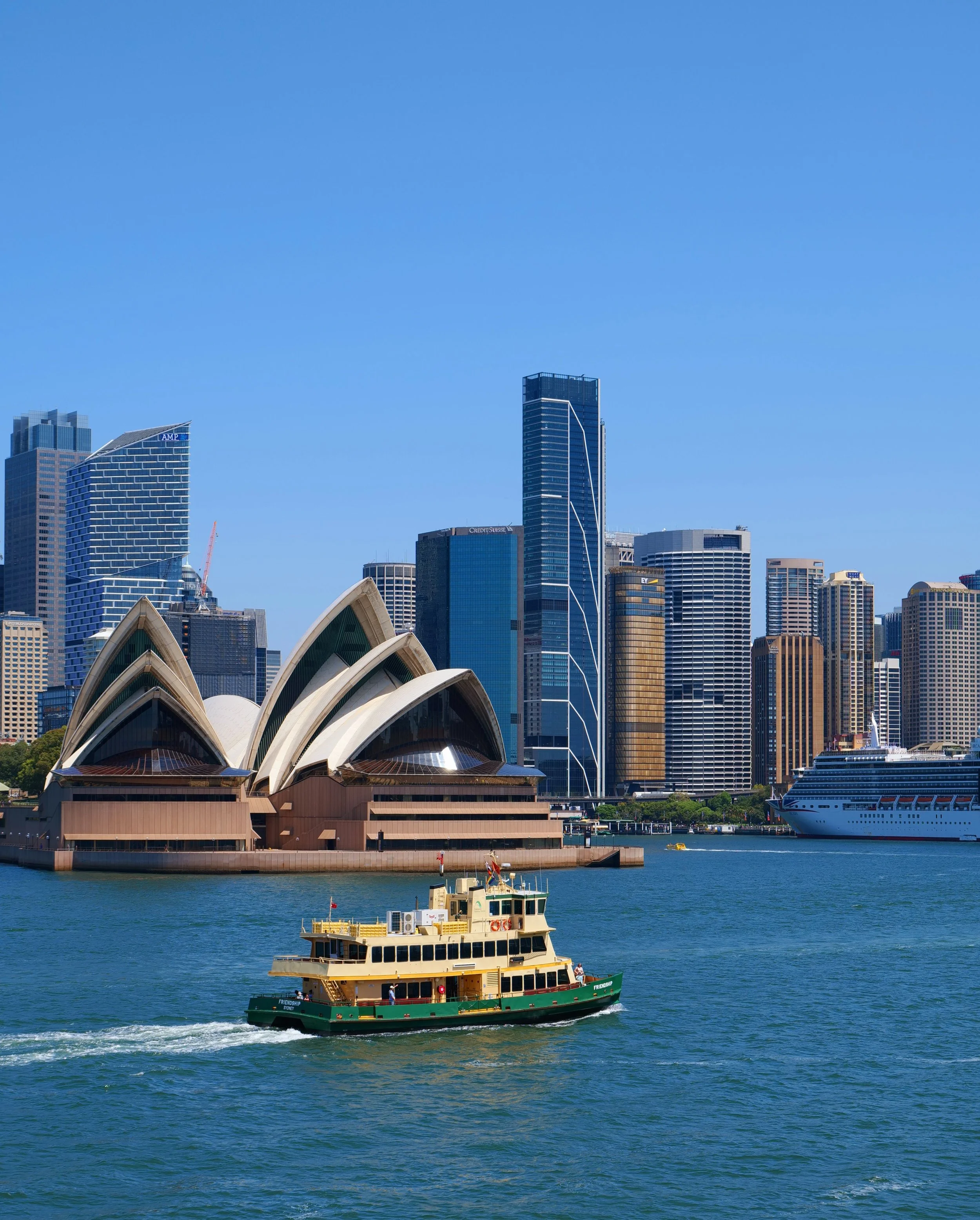 A city skyline featuring the Sydney Opera House and tall modern skyscrapers, with a ferry boat moving across the harbor in the foreground.
