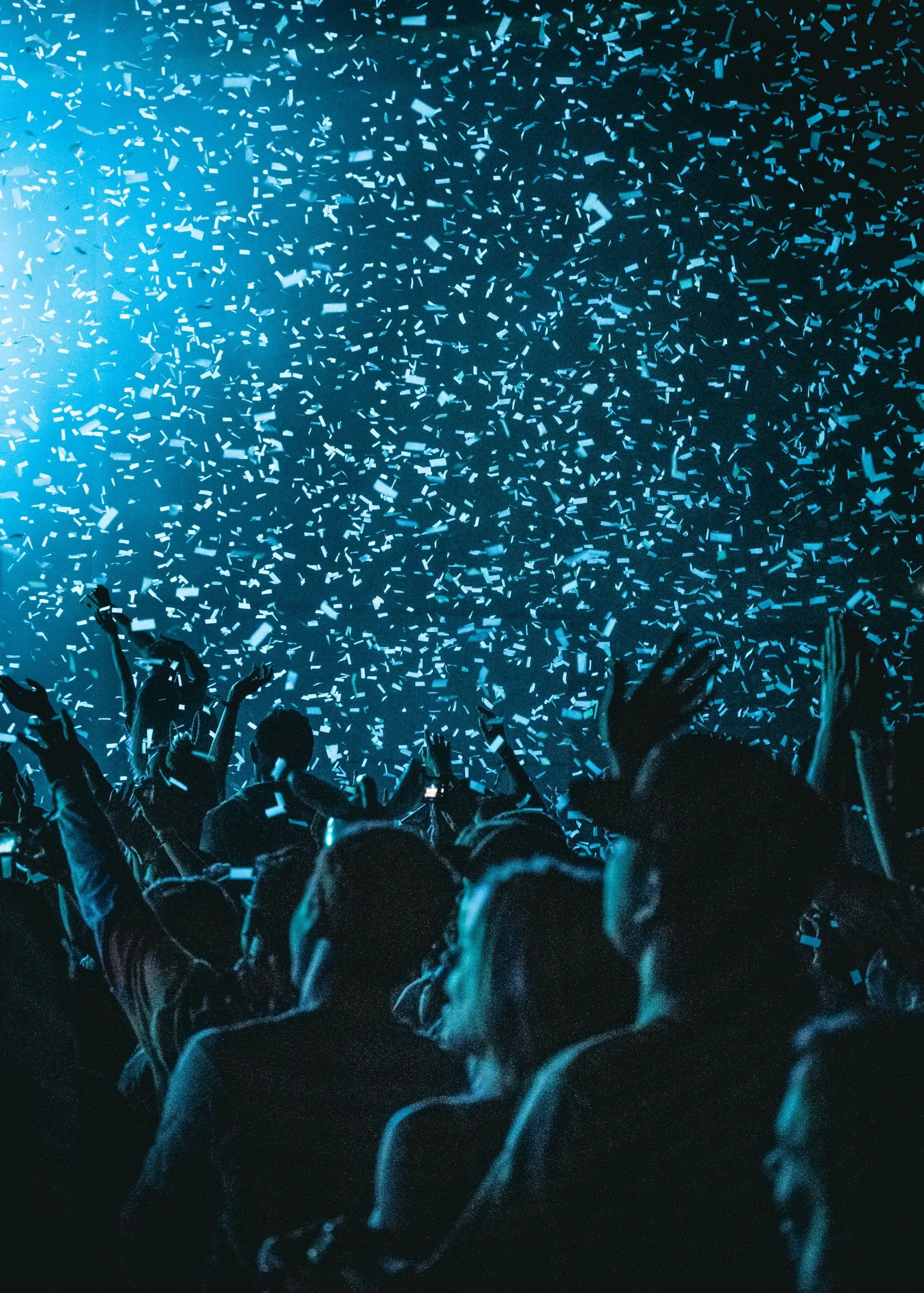 A crowd of people at a concert or event with hands raised, illuminated by blue lighting, and confetti falling from above.