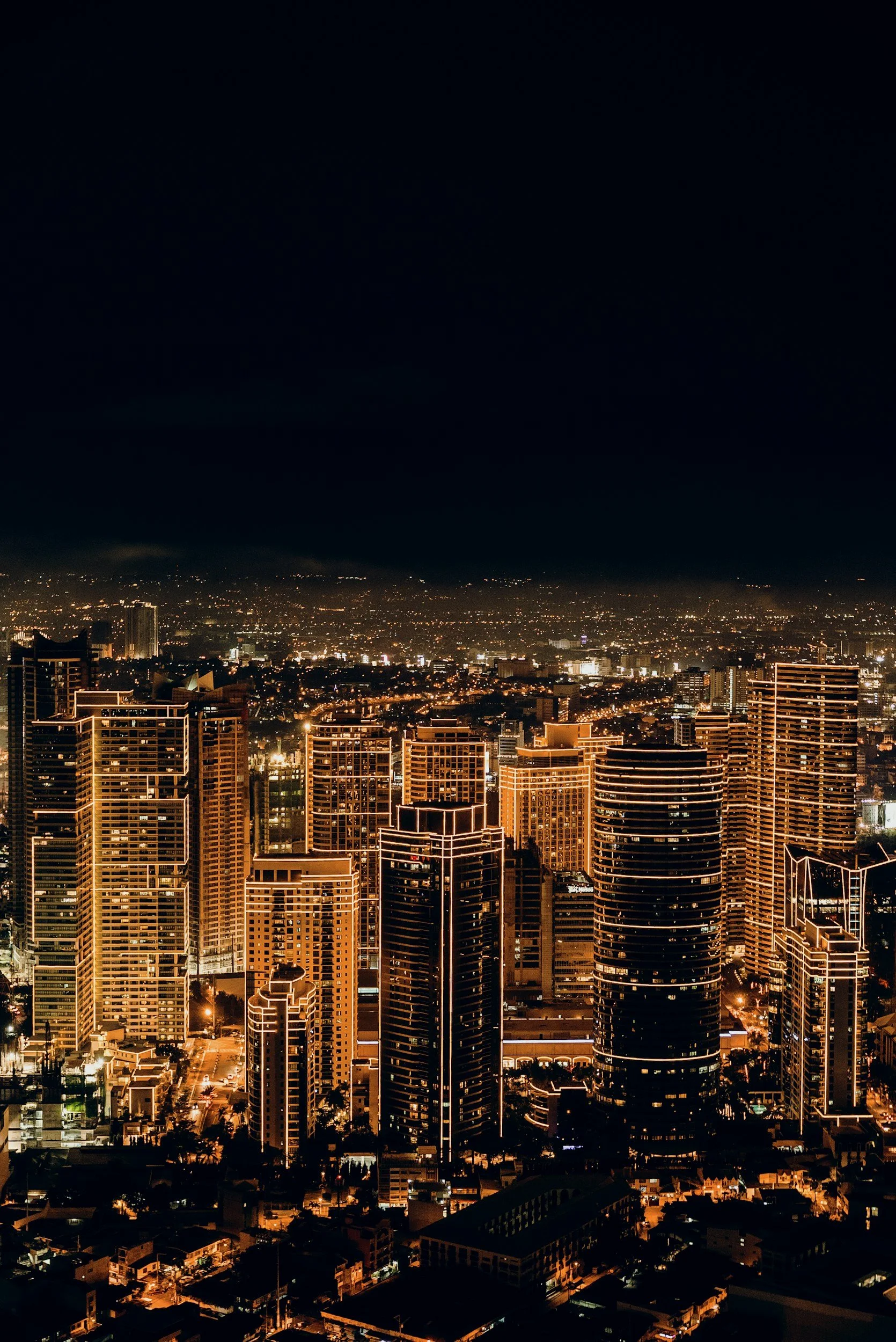 Nighttime cityscape with tall illuminated skyscrapers in the foreground and a sprawling city extending into the distance under a dark sky.