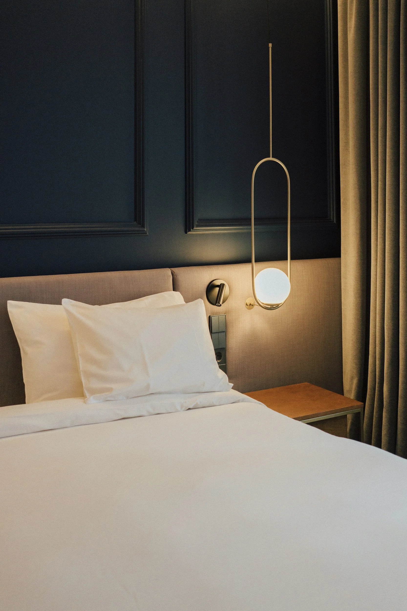 A hotel room with a bed, white pillows, and a navy blue wall with decorative molding. There is a modern pendant light and a beige curtain to the right.
