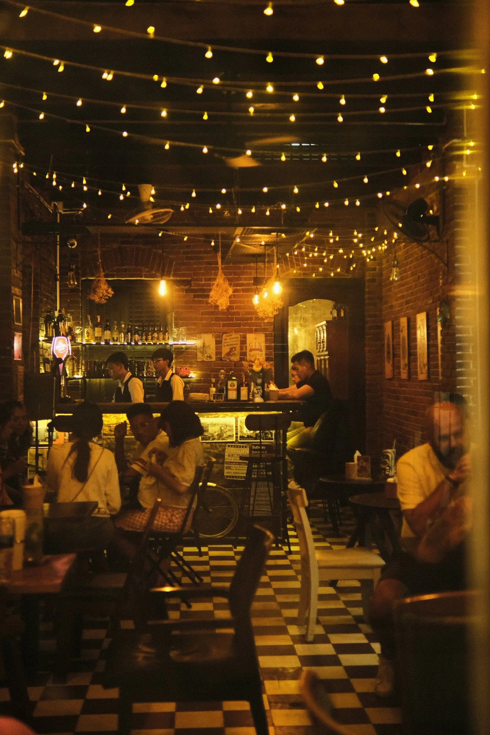 Inside a dimly lit restaurant with string lights hanging from the ceiling, a bar area with bottles displayed, and people dining and socializing, with a checkerboard floor pattern.