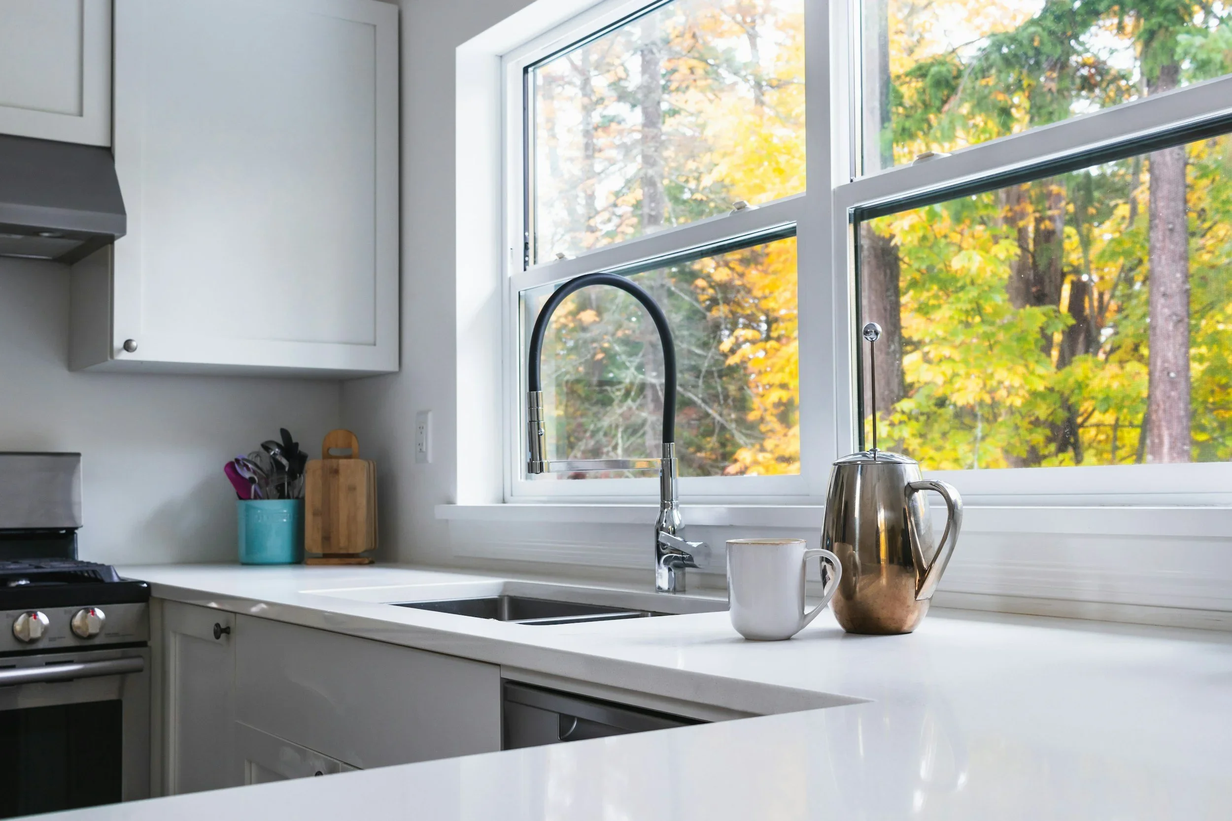 Kitchen with white cabinets and countertops, a sink near a window showing fall trees outside, a tea kettle, mug, and utensils on the counter.