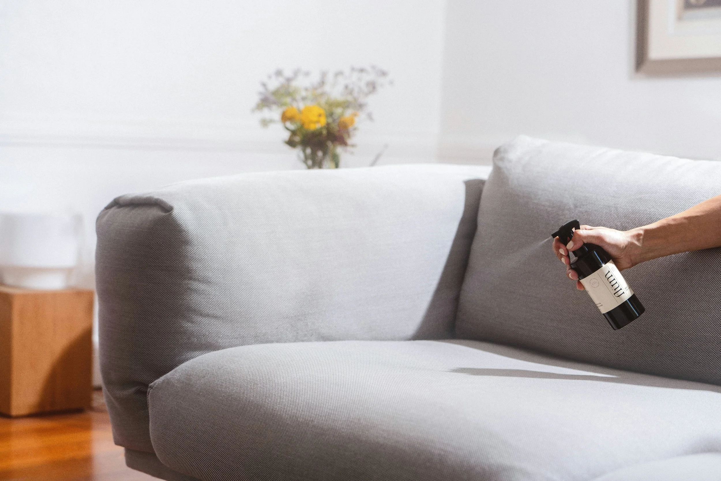 A person is spraying a fabric sofa with a cleaning or disinfectant spray. The sofa is light gray with textured fabric. There is a blurred background with a vase of yellow and purple flowers on a table.