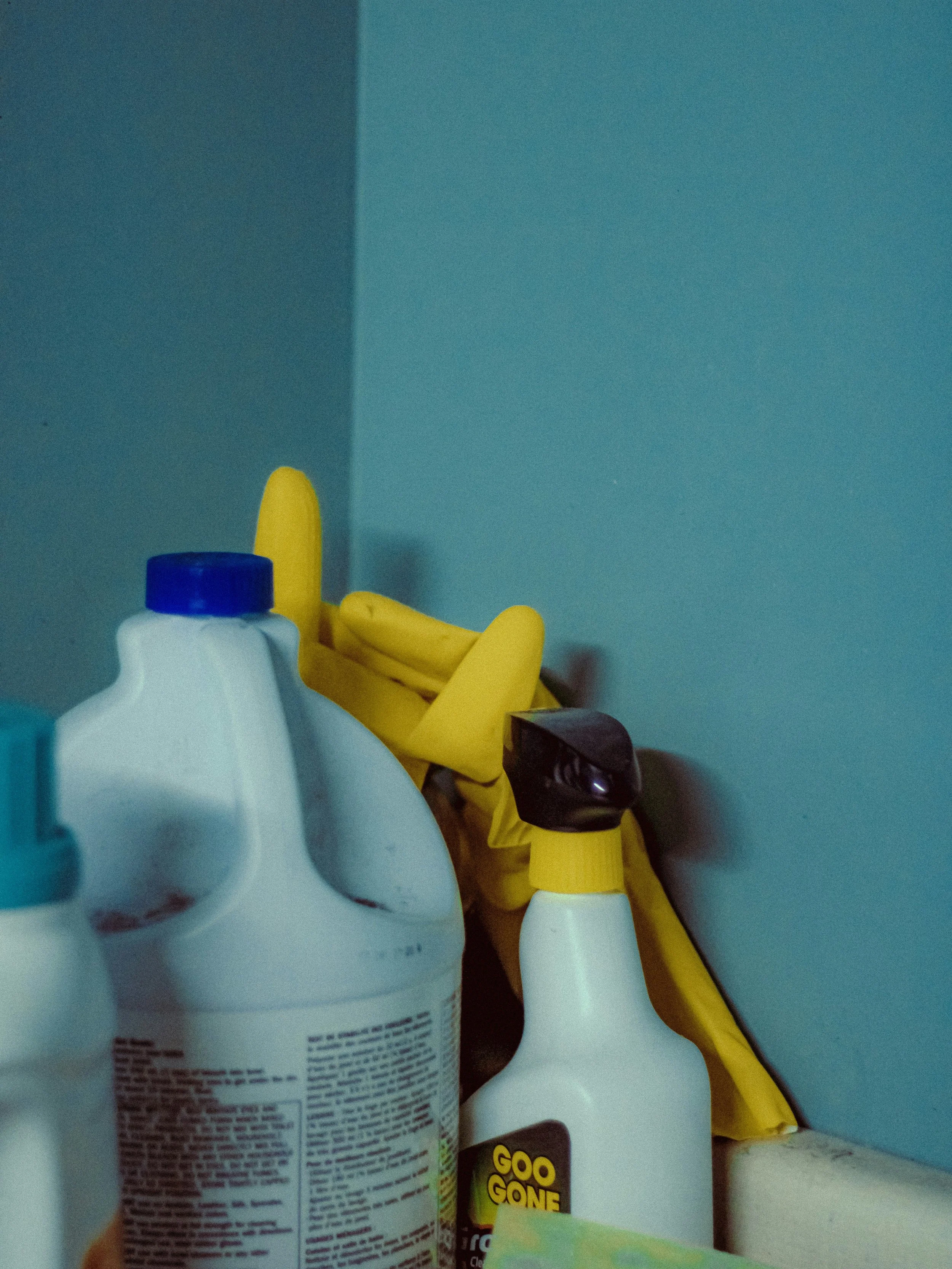 Various household cleaning products, including a spray bottle, a white bottle with a blue cap, a yellow spray bottle, and a small container with a black lid, are placed together against a plain blue wall.