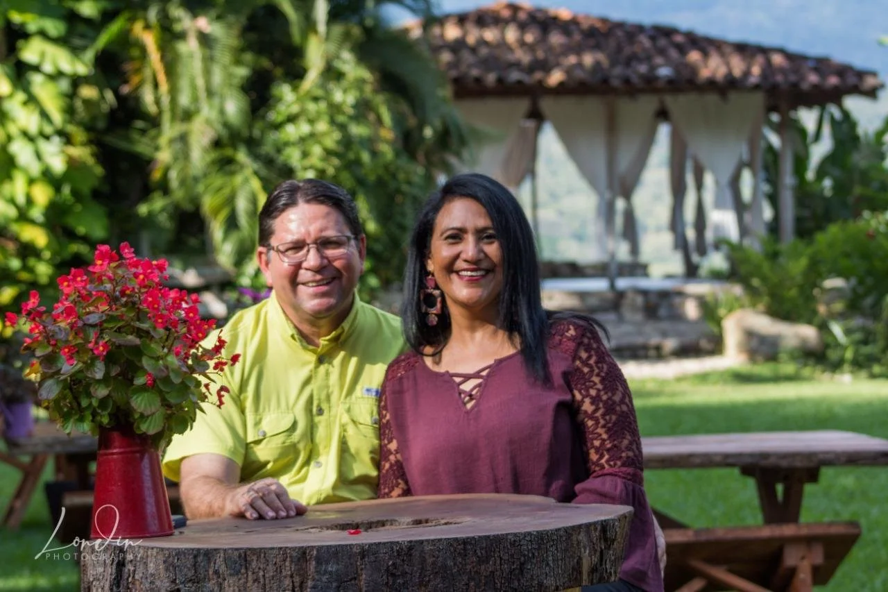 A smiling man and woman sitting together at a wooden table outdoors in a lush garden. The man is wearing glasses and a bright yellow shirt, and the woman has long black hair and is wearing a maroon blouse with lace sleeves. There is a red vase with pink flowers on the table, and a gazebo with white curtains is visible in the background.