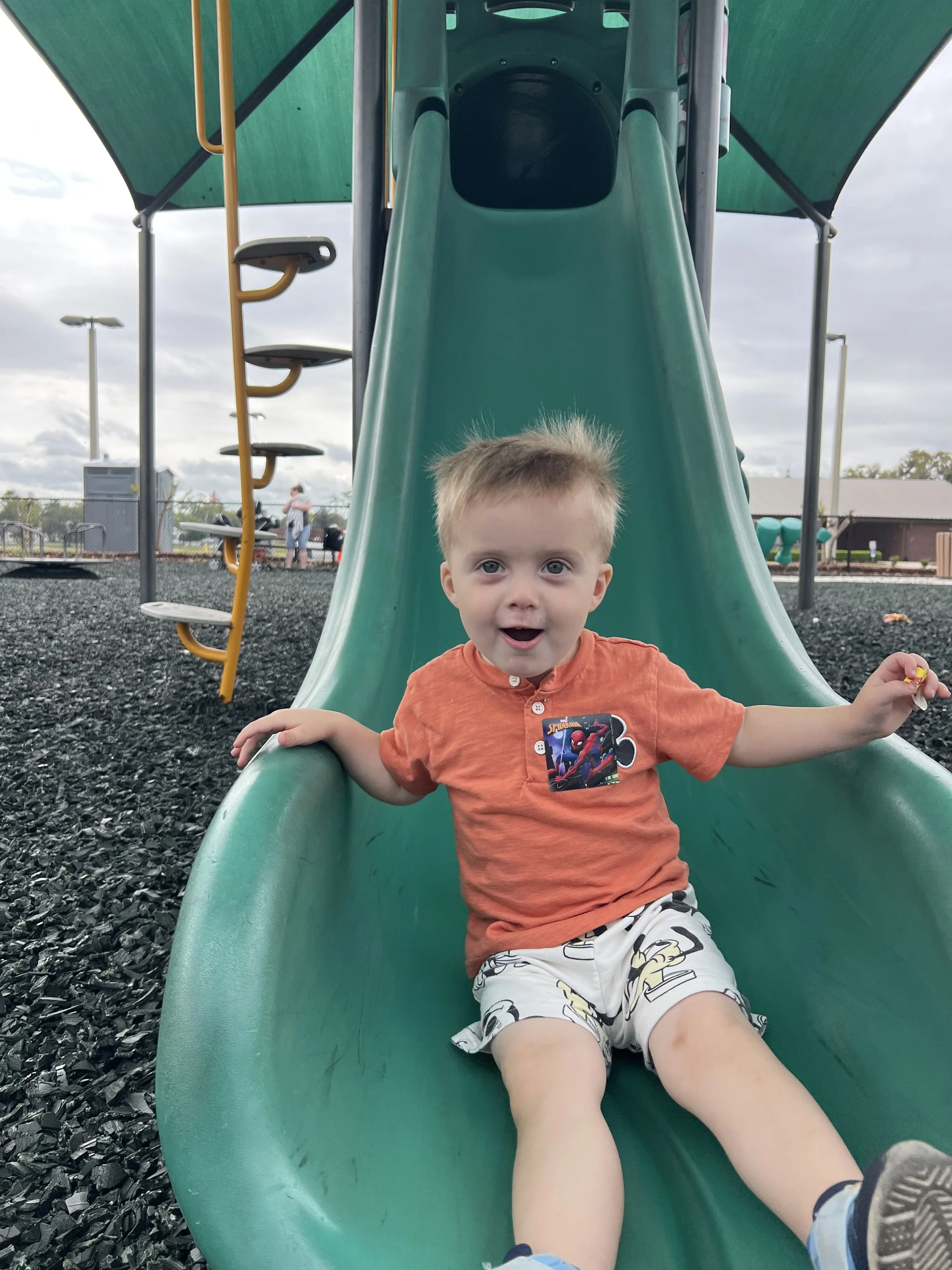 A young boy with blond hair and light skin is sitting at the bottom of a green slide on a playground, looking happy with his mouth open and arms outstretched. He is wearing an orange shirt with a Spider-Man sticker and shorts with a cartoon character.