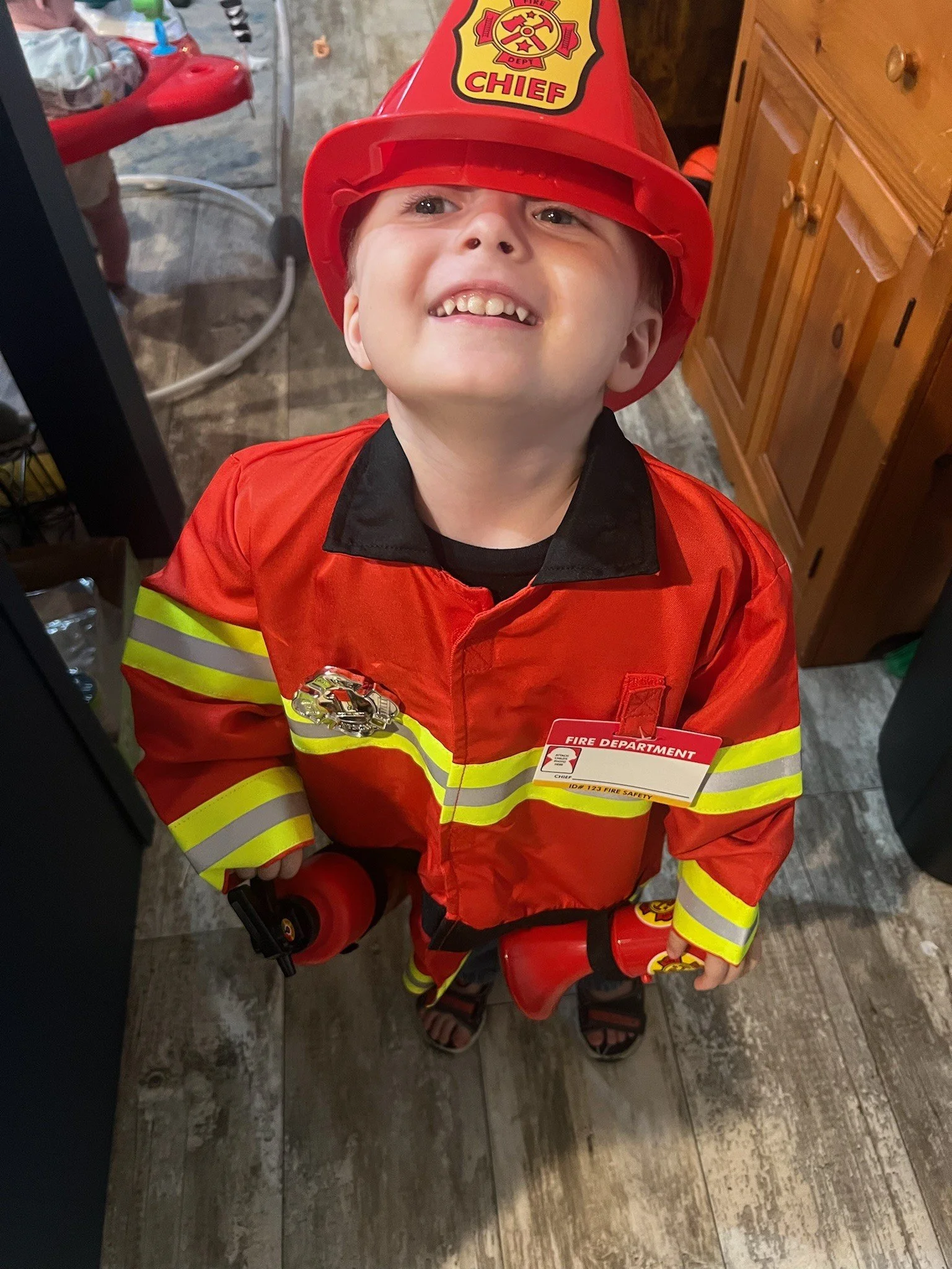 A smiling young child (Theo Maverick Harry) wearing a red firefighter costume with reflective yellow and silver stripes, a red firefighter helmet labeled “CHIEF,” and holding toy firefighting equipment while standing indoors.