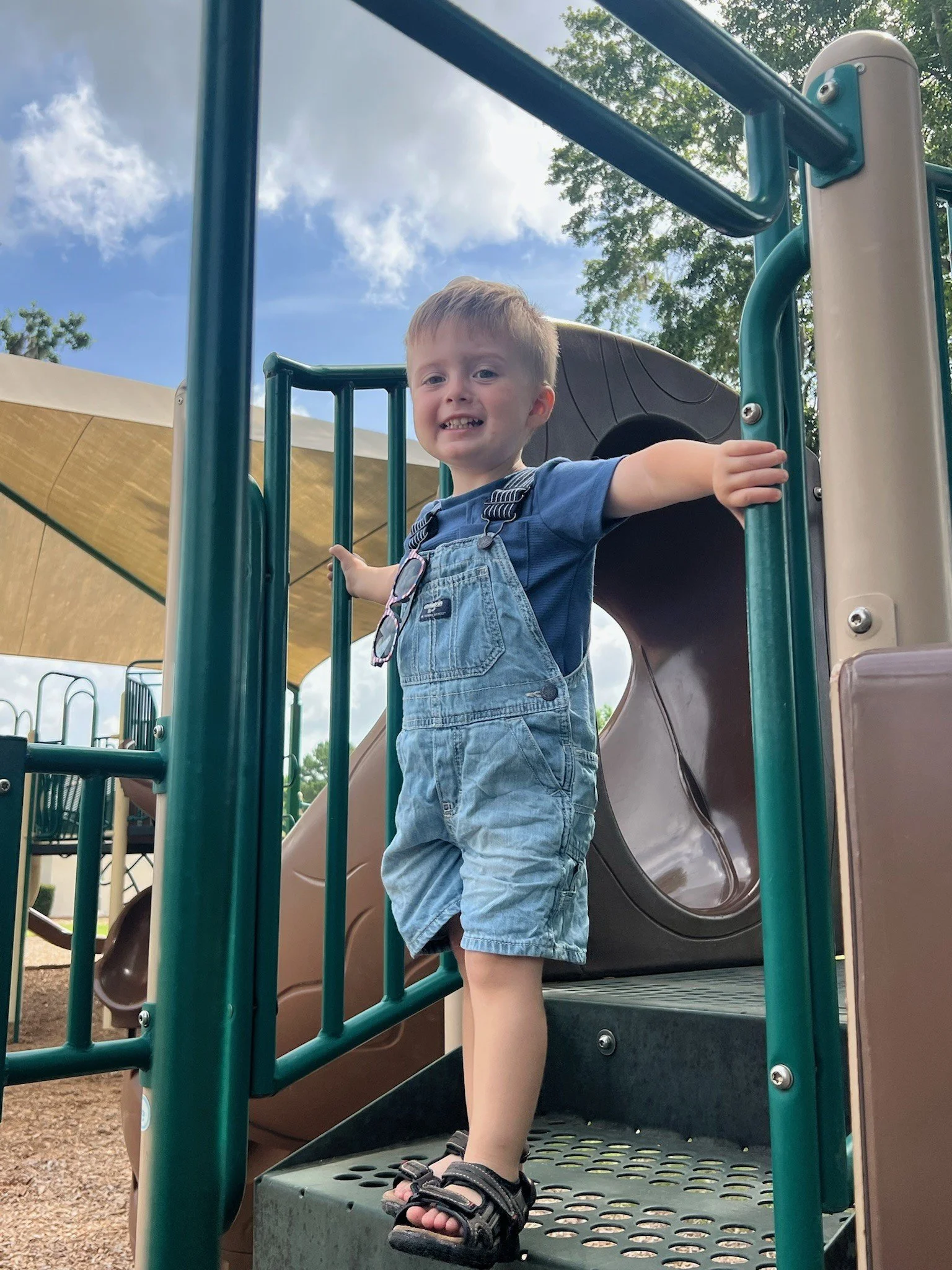 A young boy with short blond hair and wearing a blue t-shirt, denim overalls, and flip-flops, is standing on a playground platform with a slide behind him, surrounded by green playground bars and a cloudy sky above.