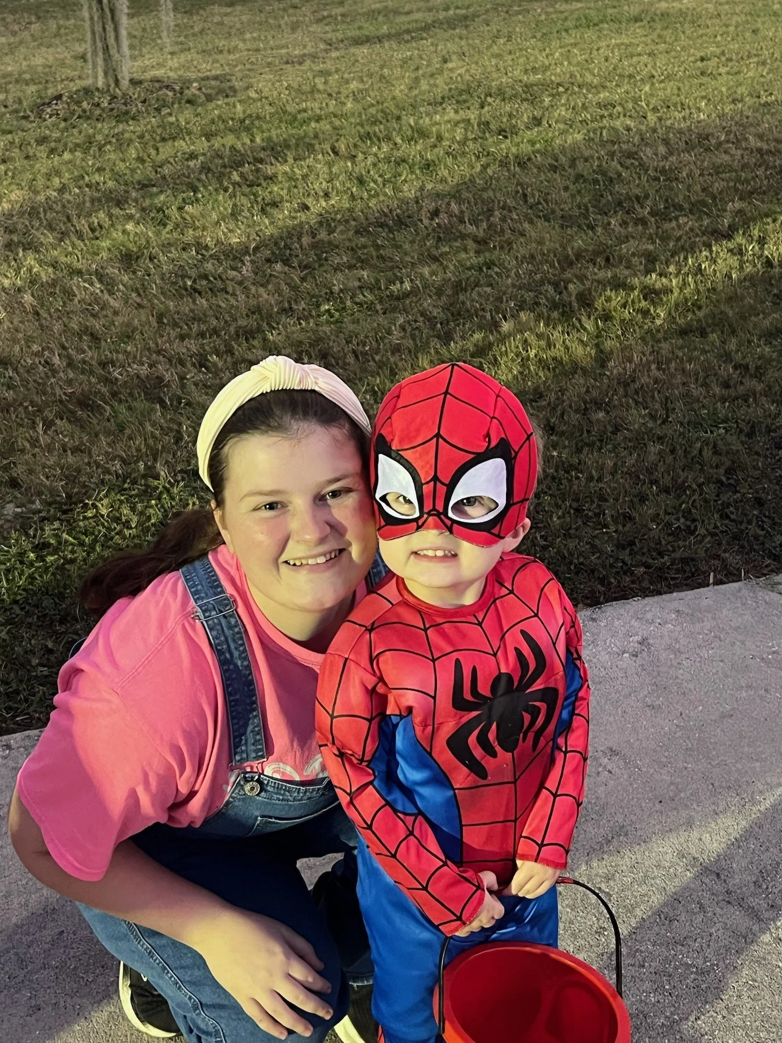 A young woman and a young boy dressed as Spider-Man posing together outdoors at night on a sidewalk with grass and a tree in the background.