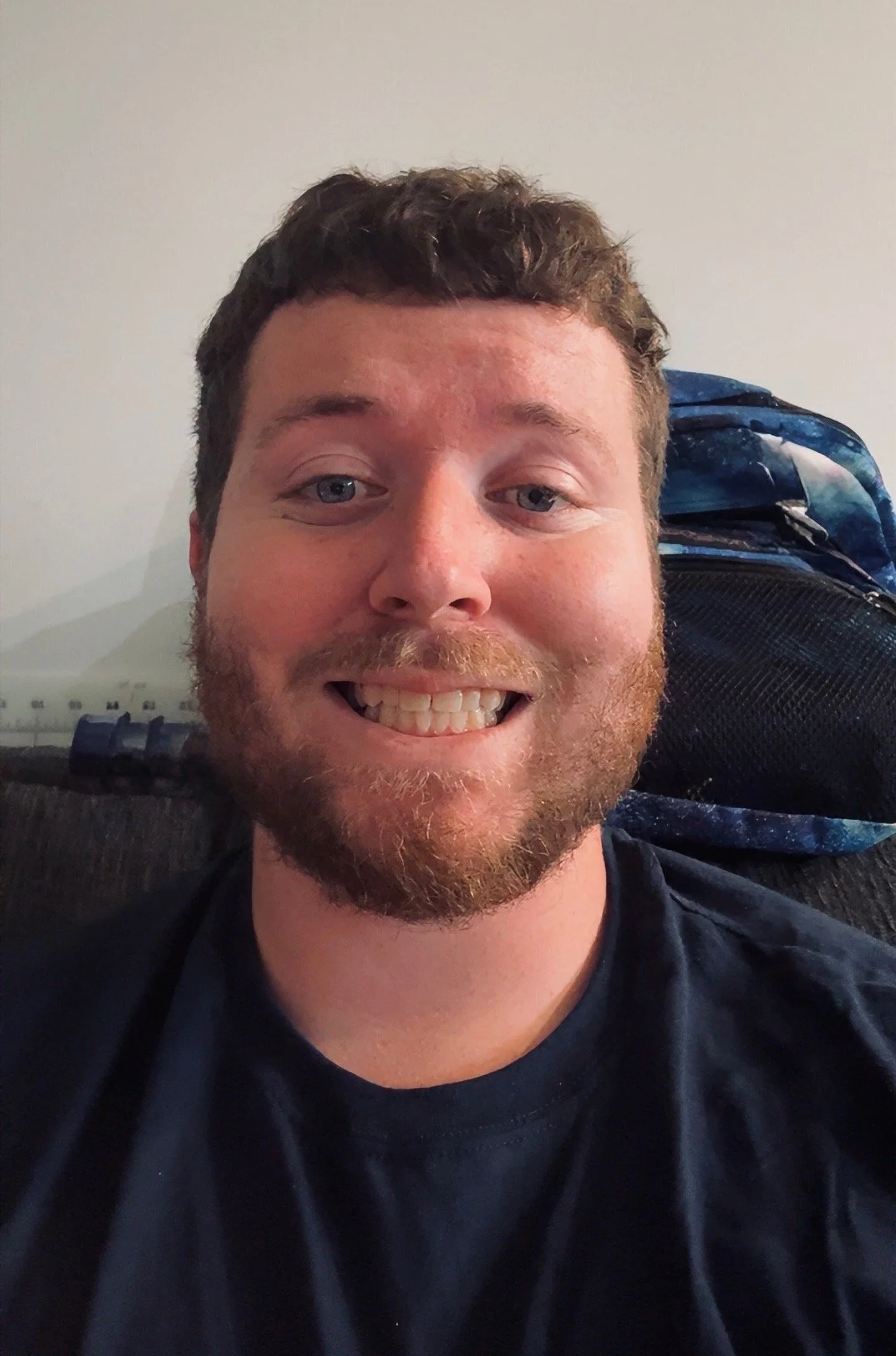 A young man with curly brown hair and a beard smiling widely, wearing a black t-shirt, and sitting indoors with a plain wall and exercise equipment in the background.