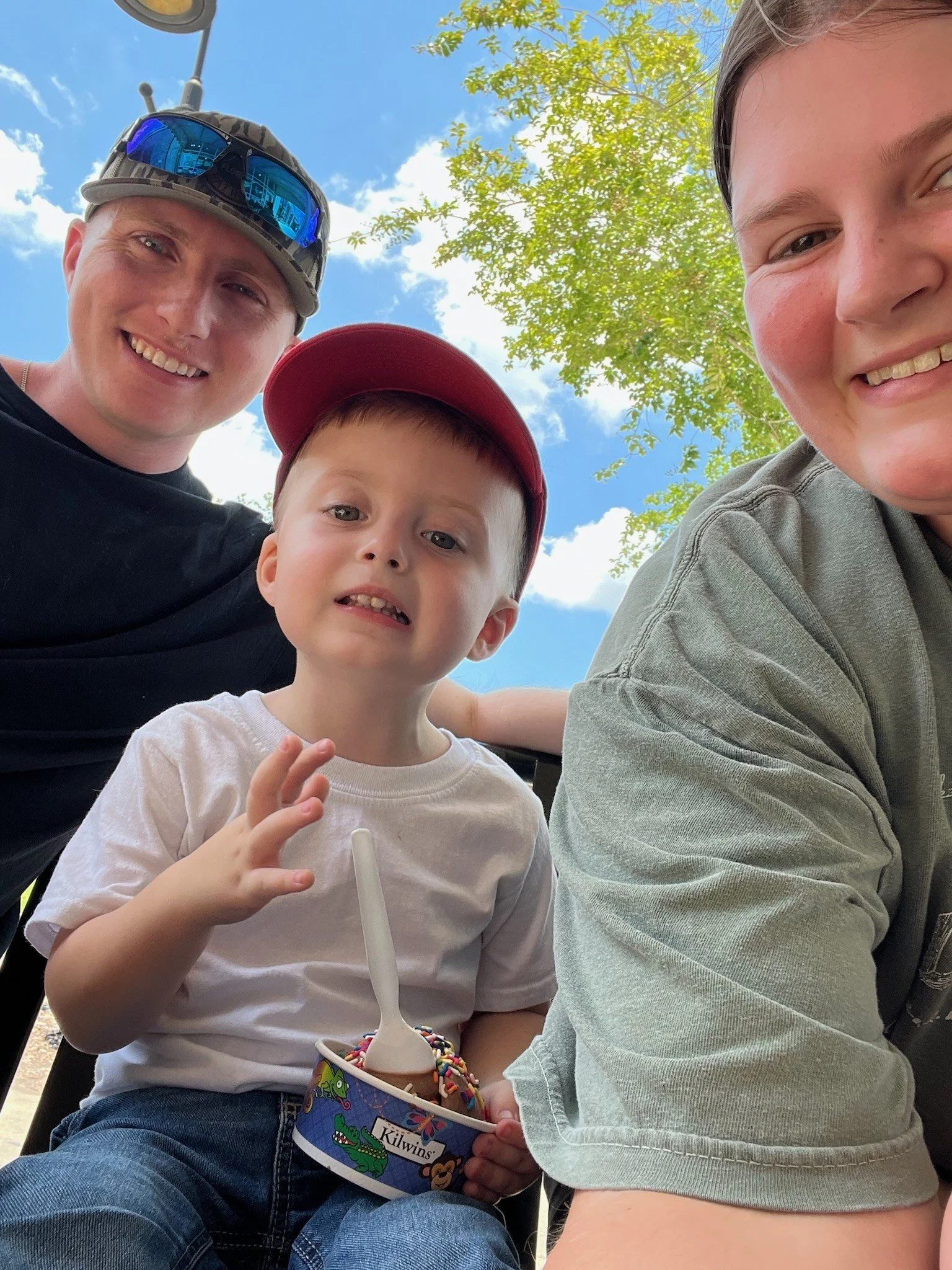 A smiling young child (Theo Maverick Harry) wearing a red baseball cap and white shirt sits outdoors with a cup of ice cream and a spoon, between two smiling adults. Trees and a bright blue sky are visible in the background.