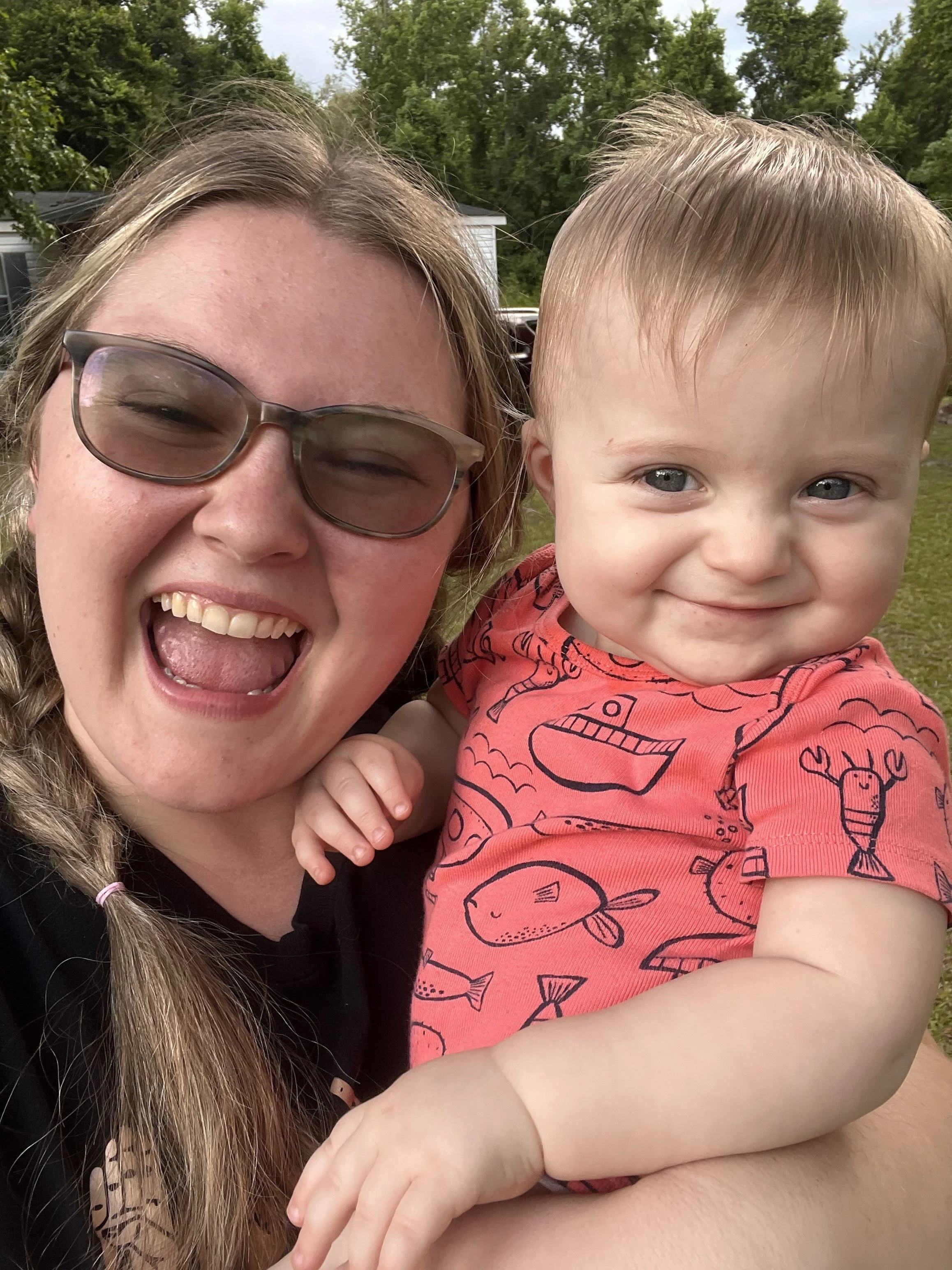 A woman with glasses and long hair smiling broadly, holding a young boy with blue eyes and short light hair, who is wearing a red shirt with fish illustrations. They are outdoors with green trees and a house in the background.