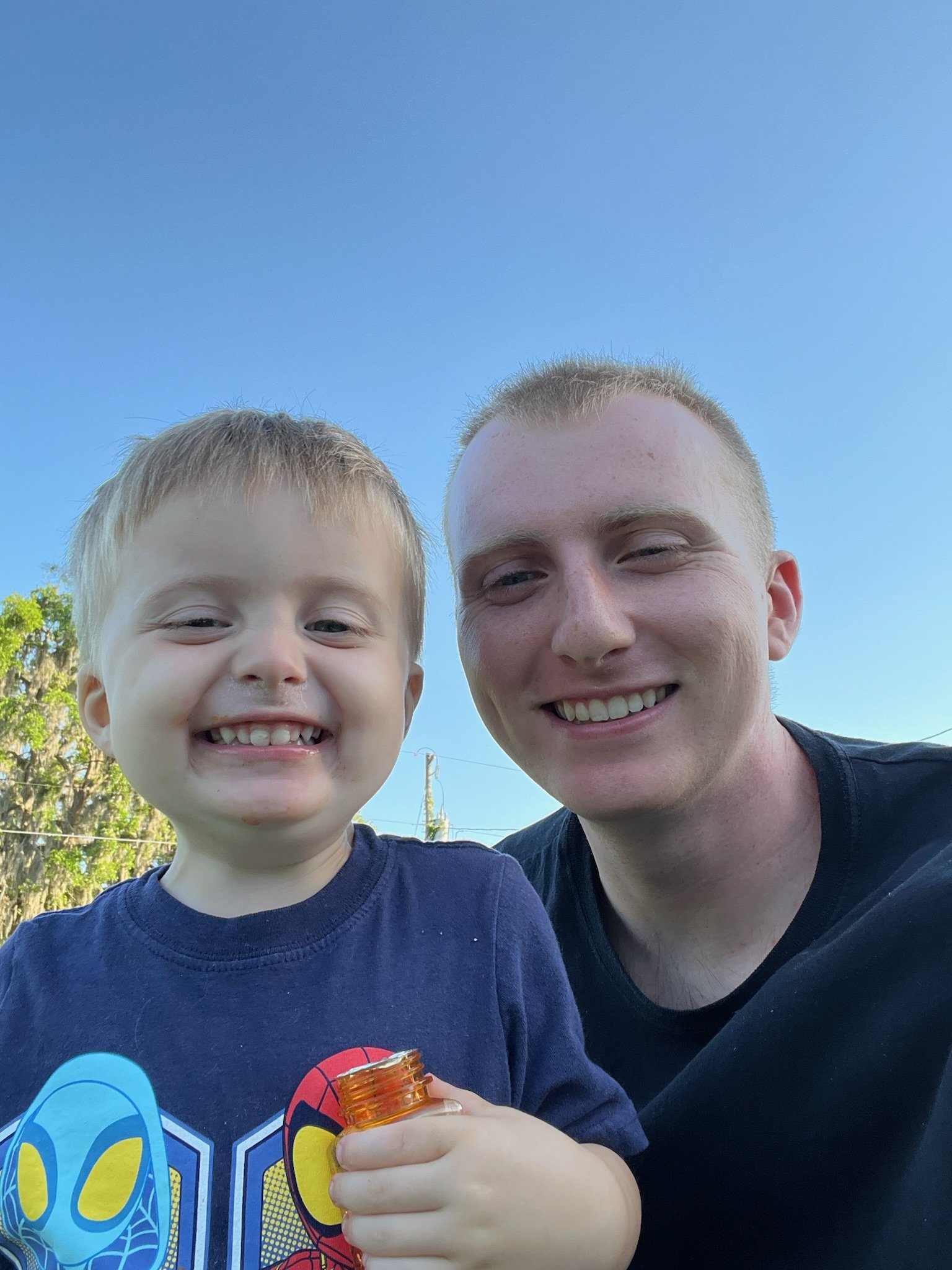 A smiling young boy and a smiling adult man taking a selfie outdoors on a sunny day. The boy is holding a small bottle of bubbles. Both are dressed casually, and the background features a bright blue sky with some trees.