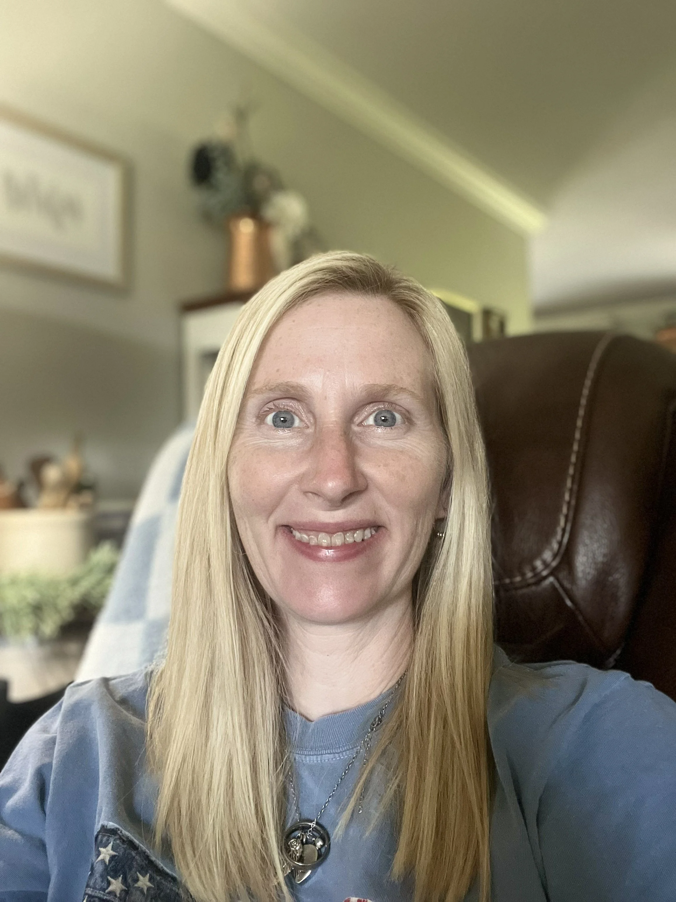 A woman with long blonde hair smiling, wearing a blue shirt with star pattern on the shoulders, a necklace with a pendant, seated on a brown leather chair, in a home setting with blurred background including a framed picture and decorative items.