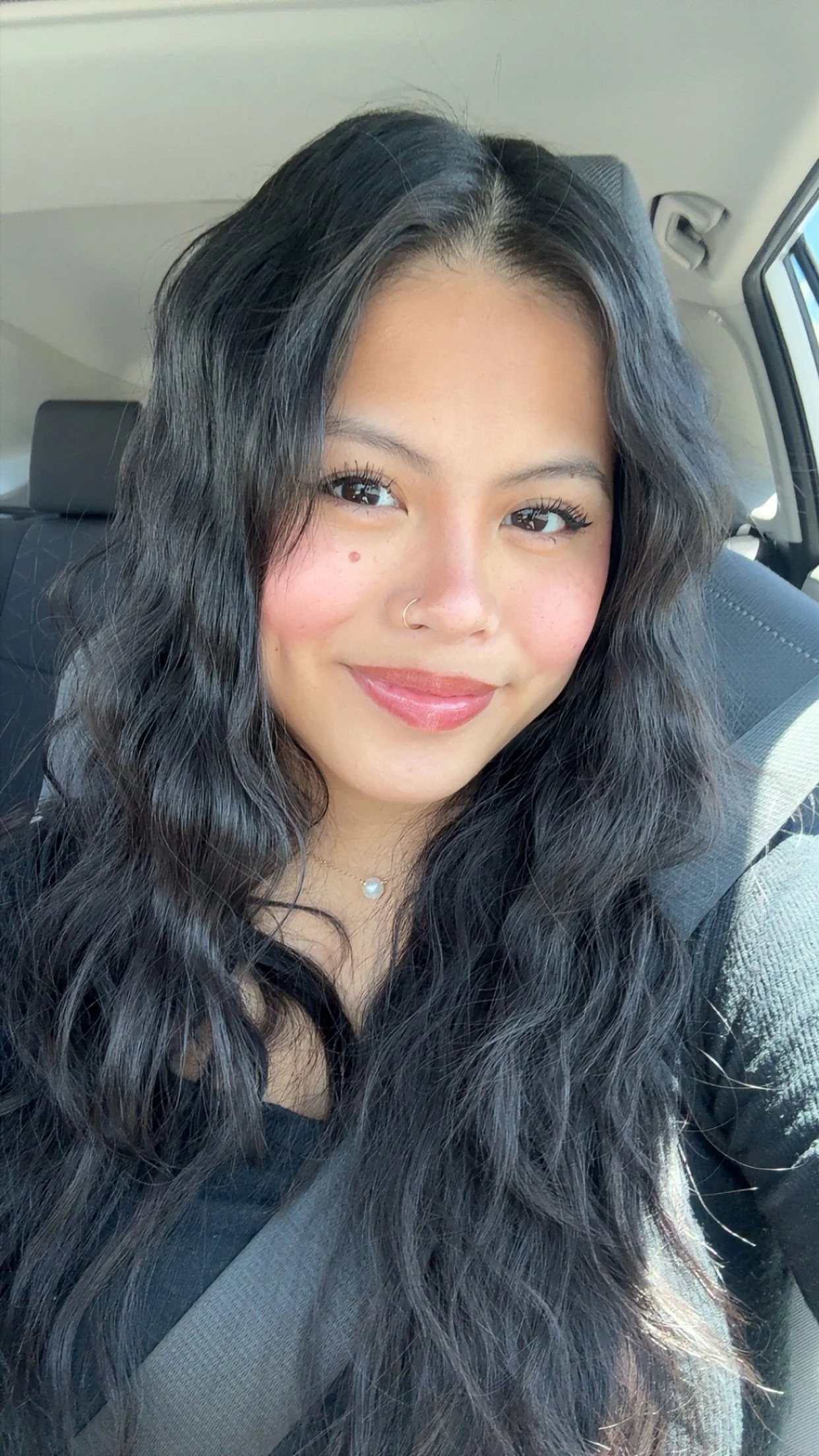 A young woman with long, wavy black hair smiling inside a car, wearing a black top, a pearl necklace, and a nose ring.