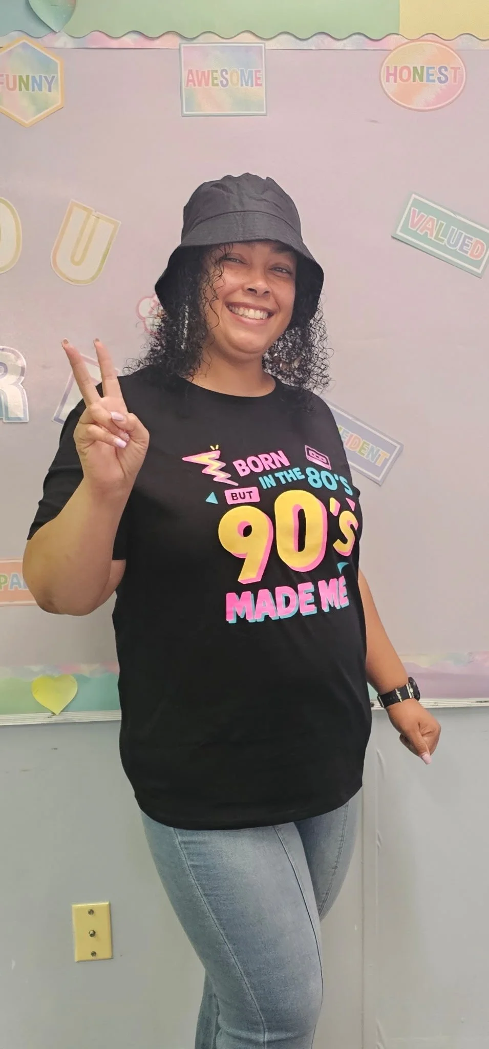 A woman with curly hair wearing a black bucket hat and a black T-shirt that reads 'Born in the 80's but made in the 90's' is smiling and making a peace sign with her right hand. She is standing in front of a pastel-colored classroom wall decorated with words like 'FUNNY,' 'AWESOME,' 'HONEST,' and 'VALUED.'