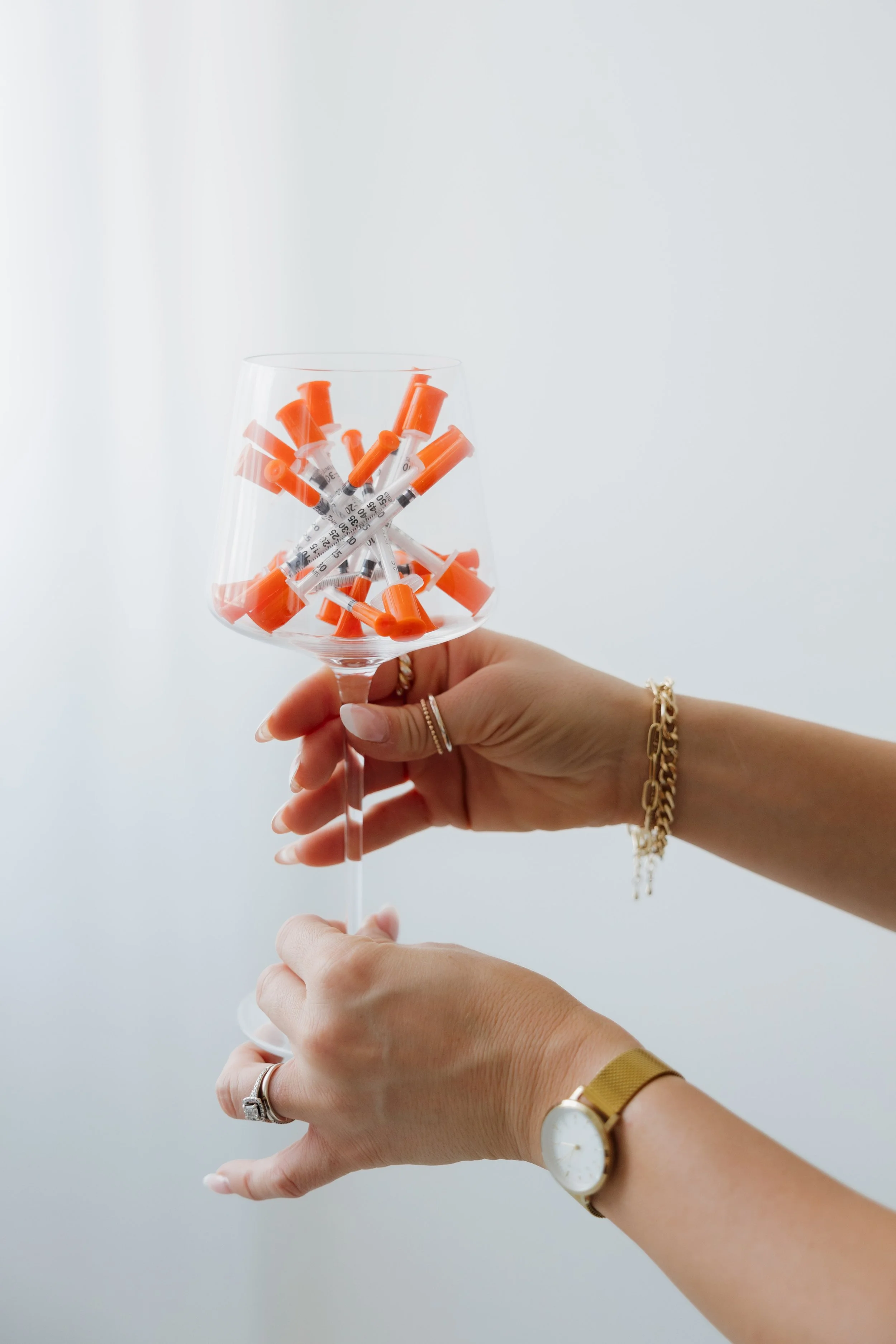 A person holding a wine glass filled with orange and white botox needles syringes against a plain light background.