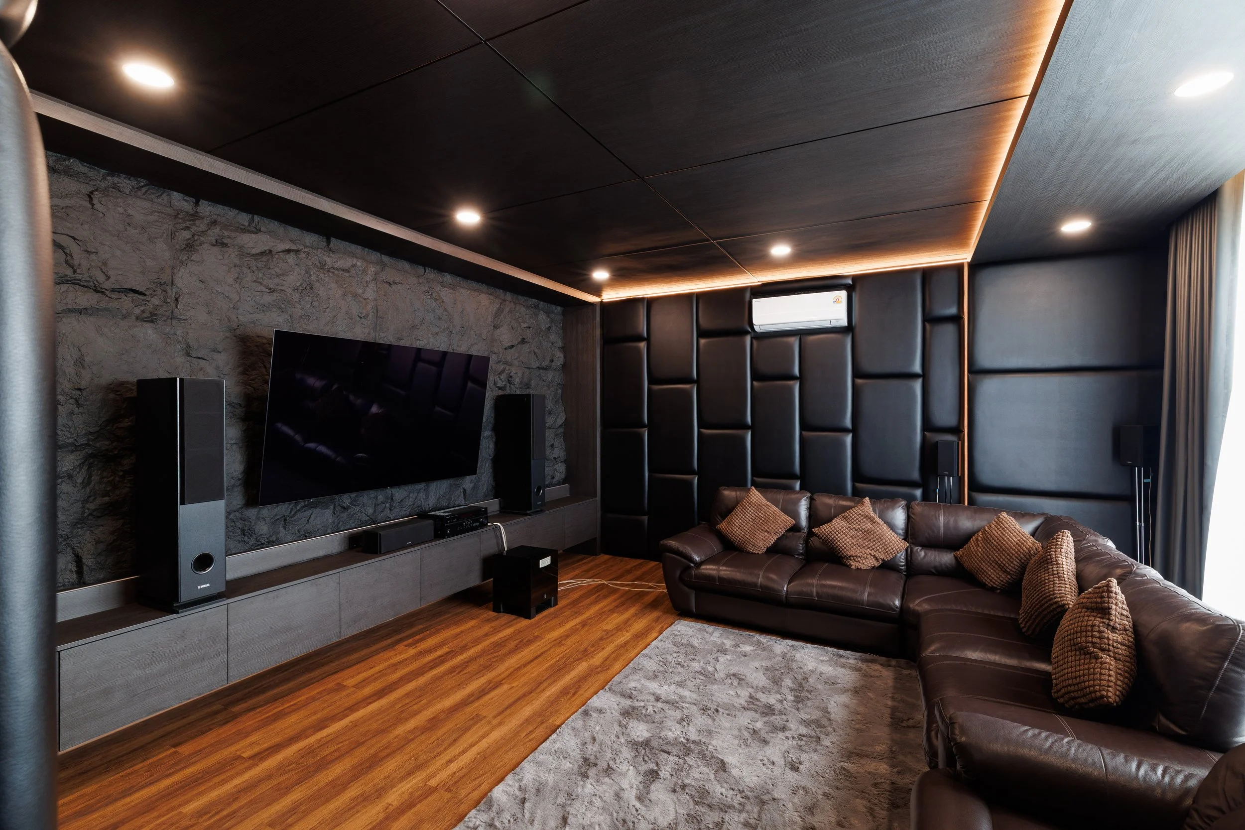Modern living room with a leather sofa, patterned cushions, a large TV mounted on a stone accent wall, and built-in speakers, hardwood floor, recessed ceiling lights, and a small window with curtains.