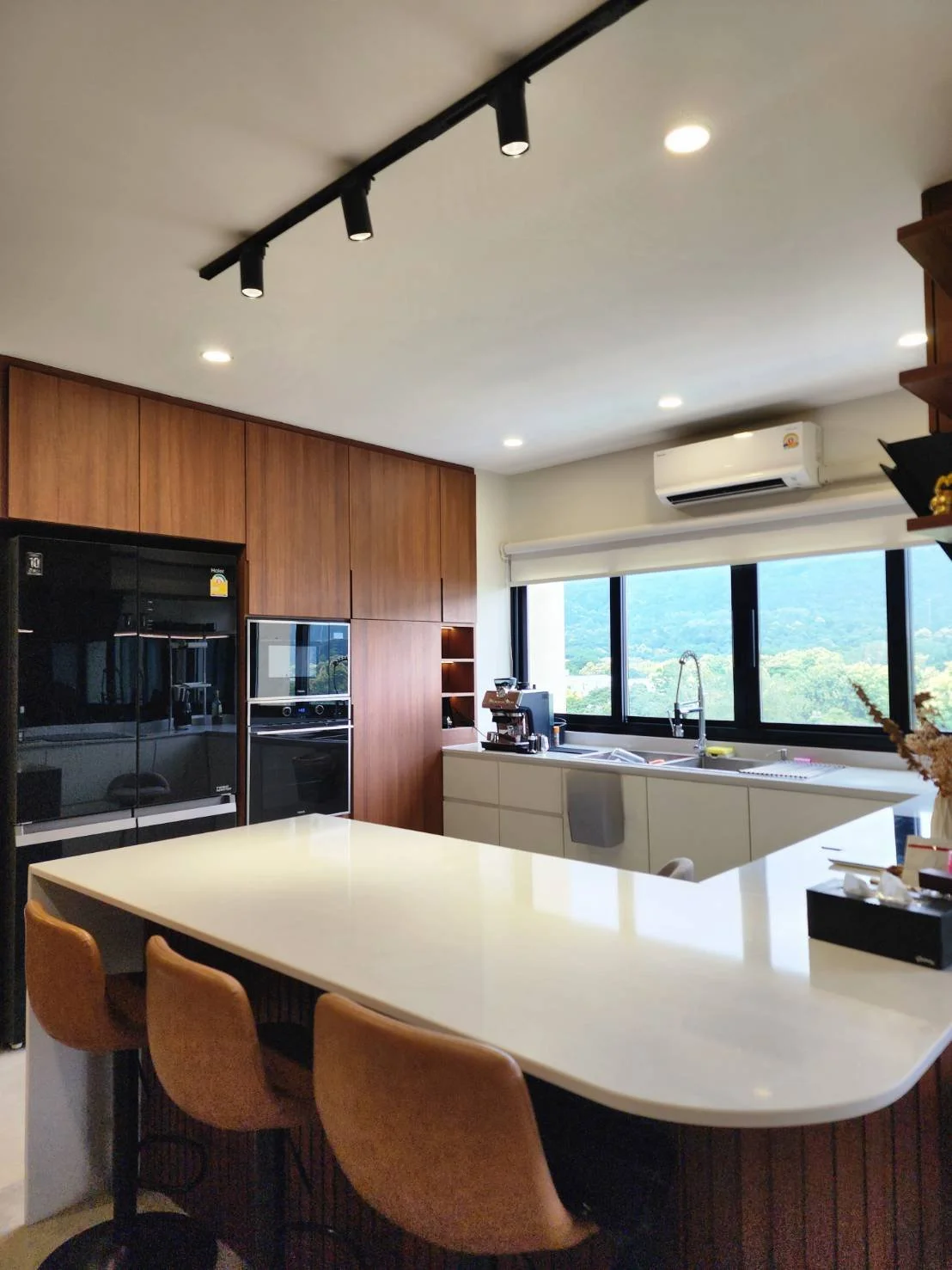 Modern kitchen with a white island counter, brown chairs, wooden cabinets, a coffee machine near the window, and a view of greenery outside.