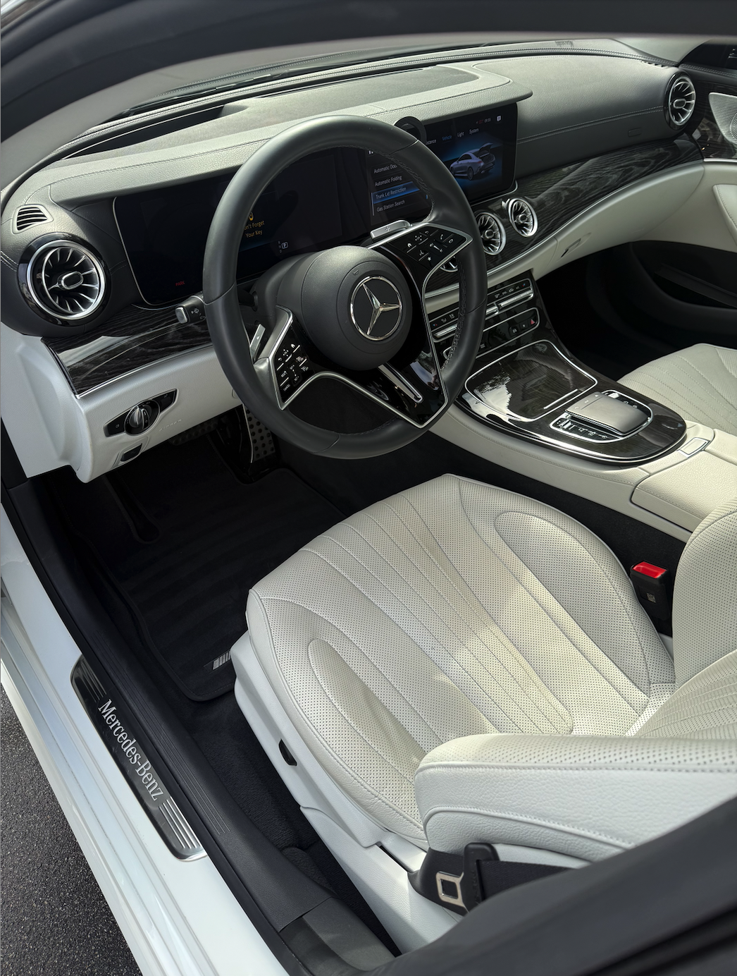 Interior view of a luxury Mercedes-Benz car with a white leather seat, black steering wheel with Mercedes logo, digital dashboard, and center console with controls and screens.