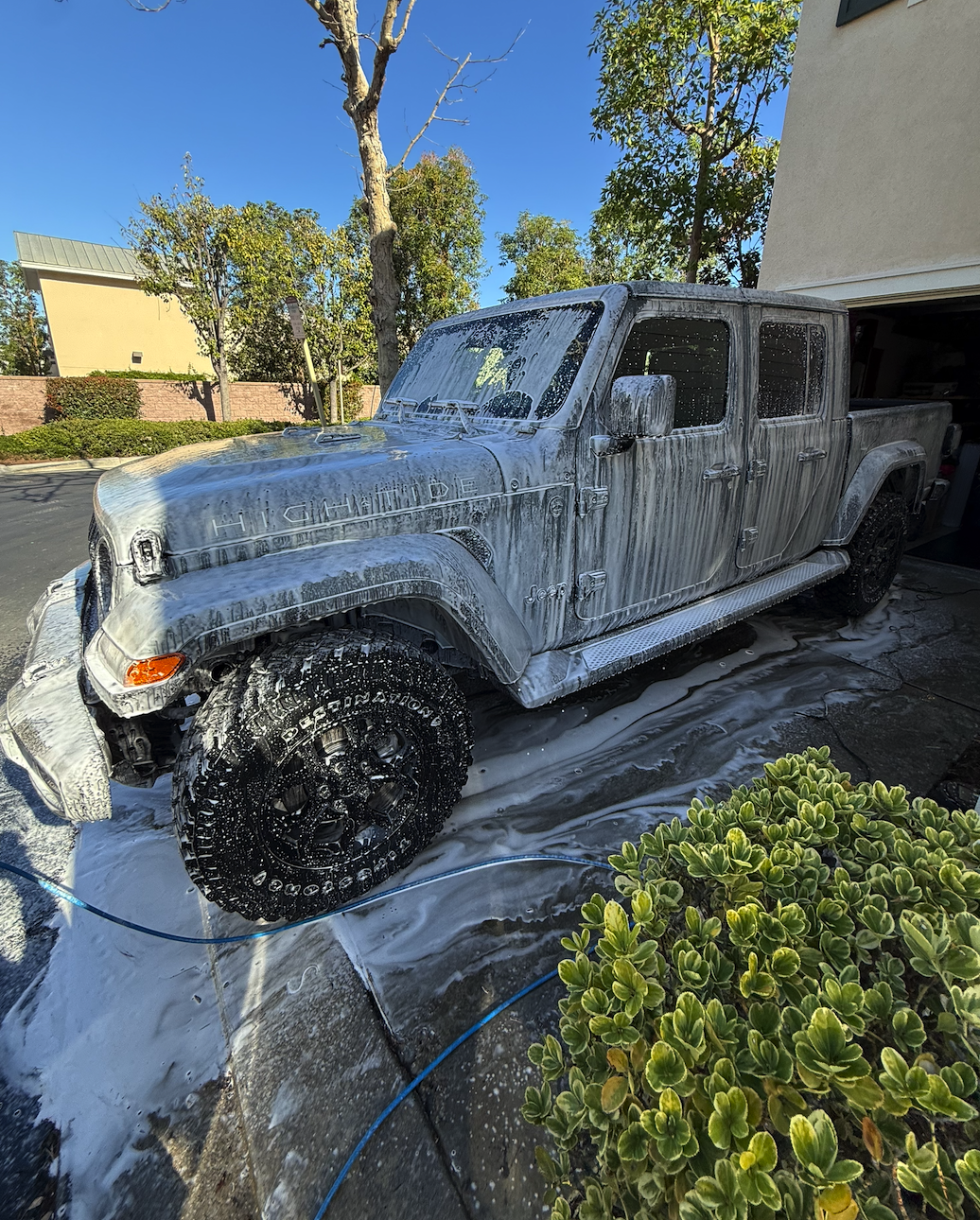 A gray Jeep Gladiator truck covered in white soap suds, being washed outside in a driveway. The Ford F-150 badge is visible on the hood, indicating it is a Ford truck.