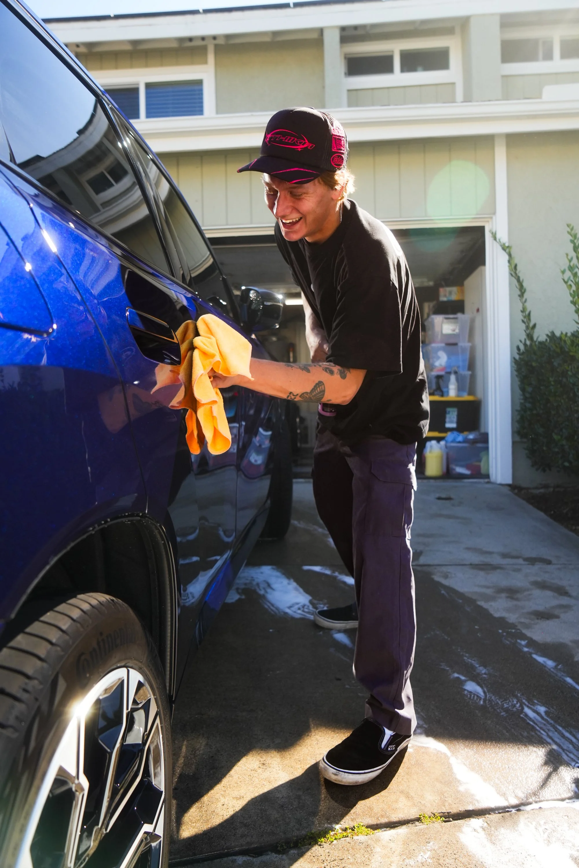 A young man is washing a blue car in a driveway, using a yellow cloth. He is wearing a black cap with pink details, a black t-shirt, gray pants, and black and white sneakers. His tattoos are visible on his left arm, and he is smiling as he leans under the car's side mirror.