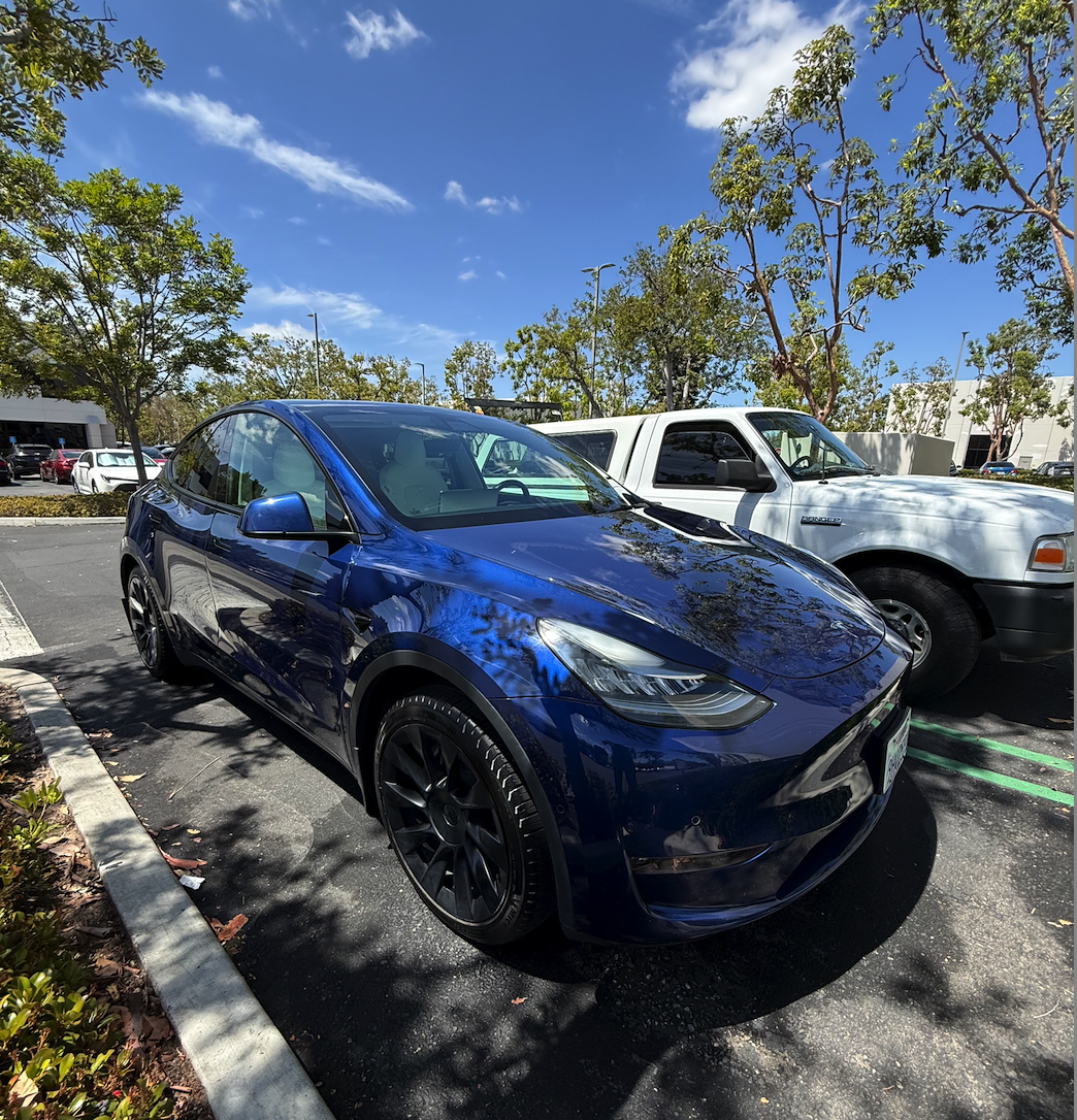 Blue Tesla Model Y parked in a parking lot beside a white pickup truck, with trees and a building in the background under a partly cloudy sky.