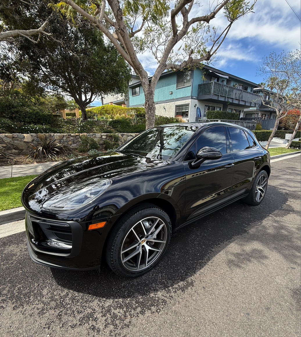 A black Porsche Macan parked on a residential street with houses, trees, and a partly cloudy sky in the background.