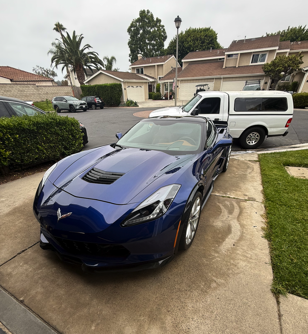 A blue Chevrolet Corvette sports car parked on a driveway next to a white van and other vehicles in a suburban neighborhood.