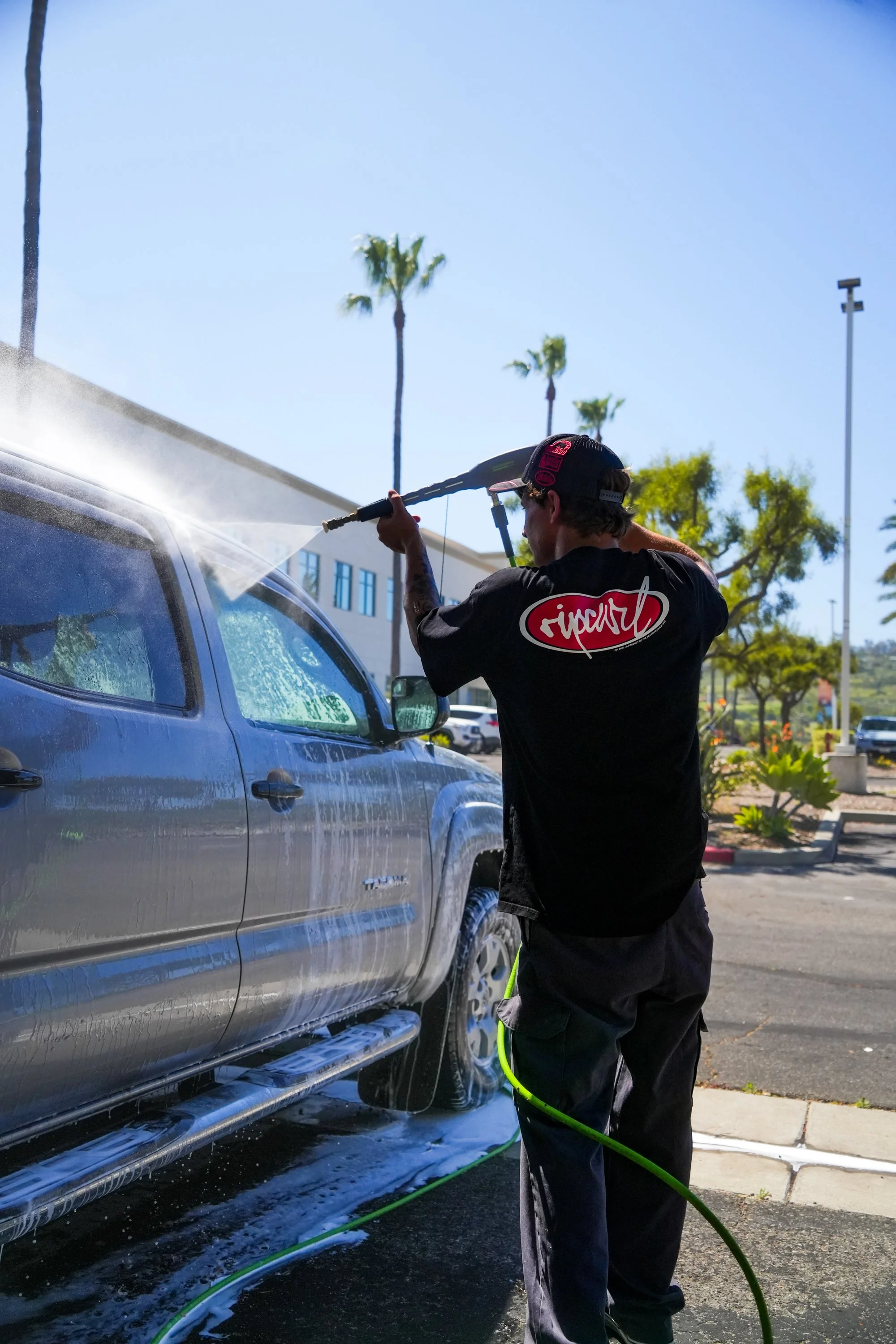 A man cleaning a silver pickup truck with a pressure washer in a parking lot on a sunny day, with palm trees in the background.