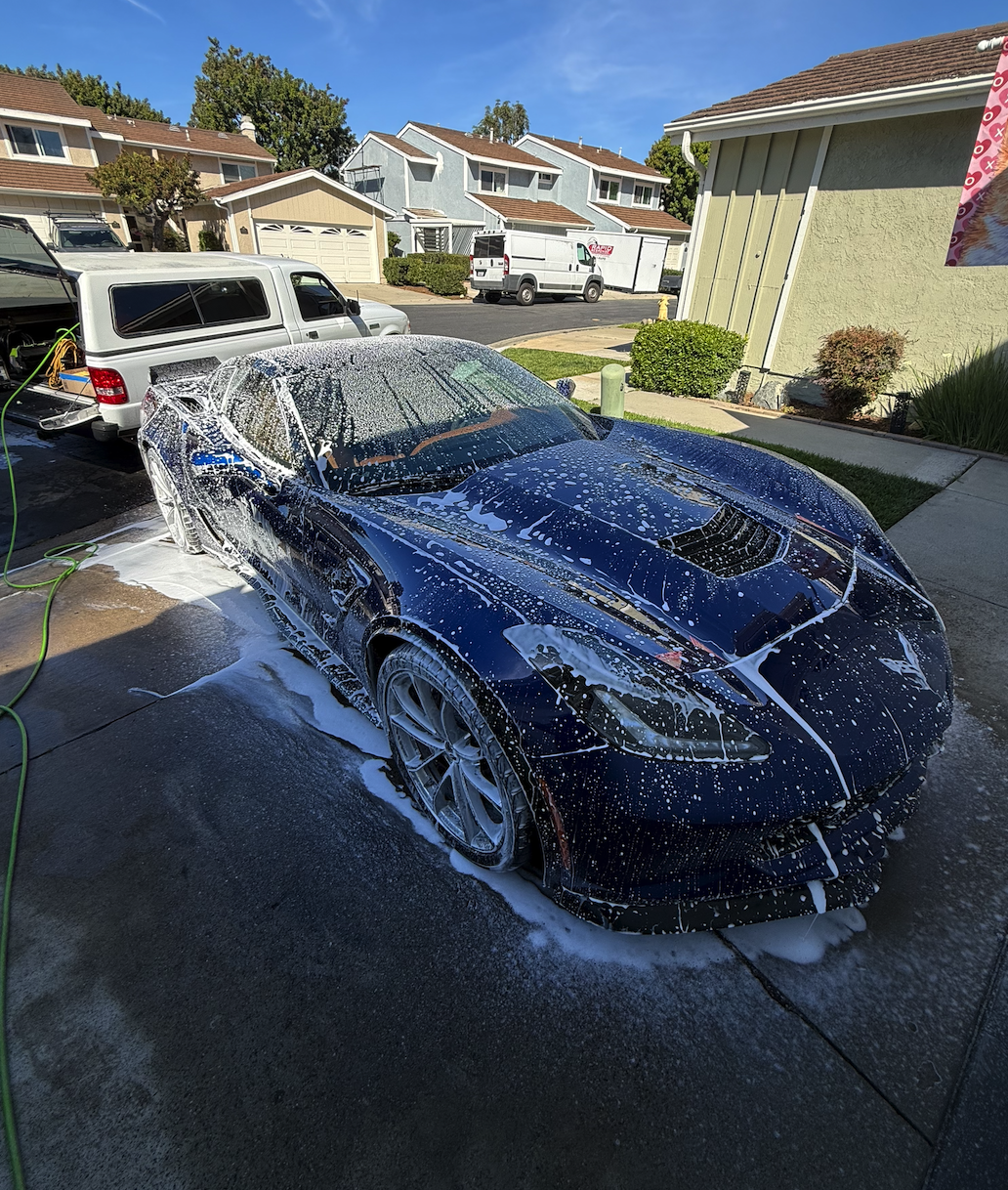 A black sports car being washed with soap and water, creating foam and suds on the vehicle's surface, parked on a driveway in front of a suburban house on a sunny day.