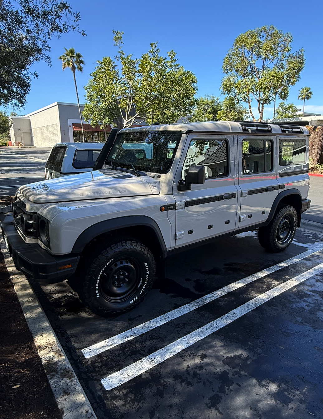 A silver Land Rover Defender parked in a parking lot under a clear blue sky, with trees and a building in the background.