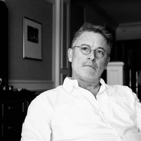 A black and white photo of a man with glasses, short hair, and a beard sitting in a room, wearing a white shirt.