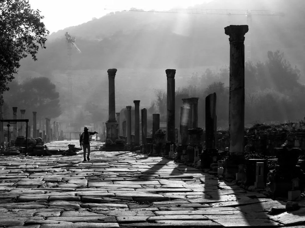 A person walking through ancient ruins with stone columns, during sunrise or sunset, with a mountain in the background and fog or haze surrounding the area.