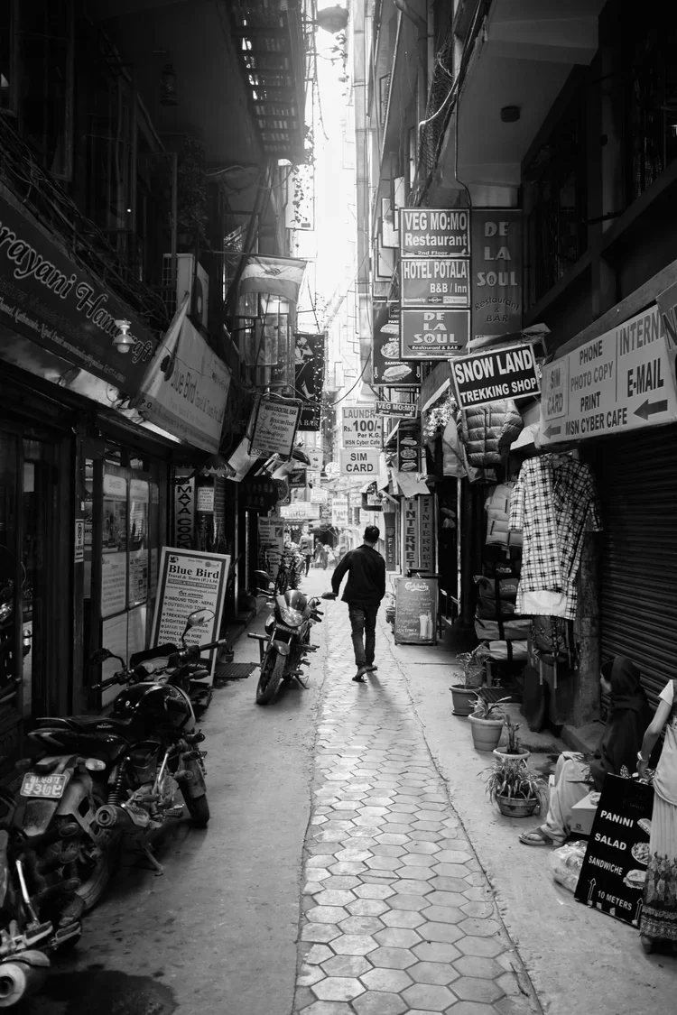 A narrow city alley with shops on both sides, motorcycles parked along the curb, and a person walking away in the distance.