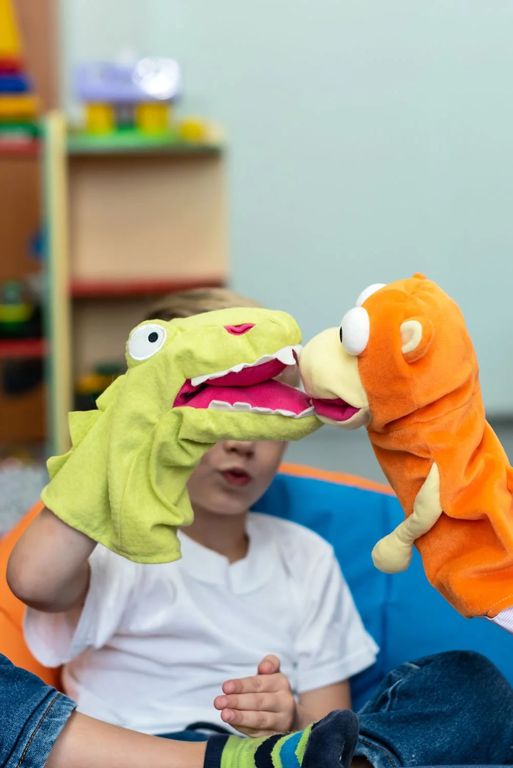 Child playing with two colorful hand puppets during a Innate Play Therapy Session, a green one resembling a dinosaur and an orange one resembling a monkey, in a playroom.