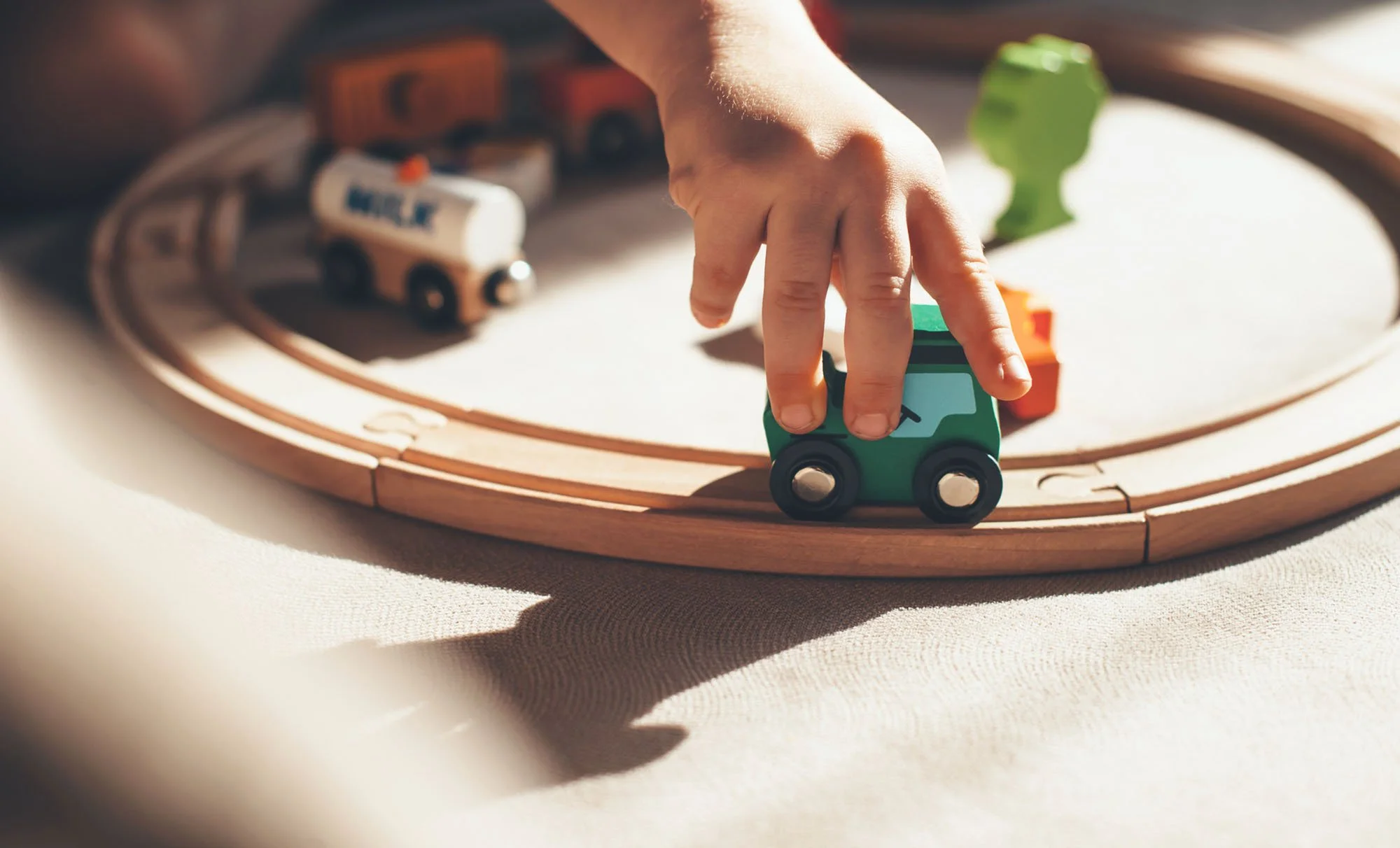 Child's hand moving a small green toy train on a wooden train track with other toy vehicles in the background.