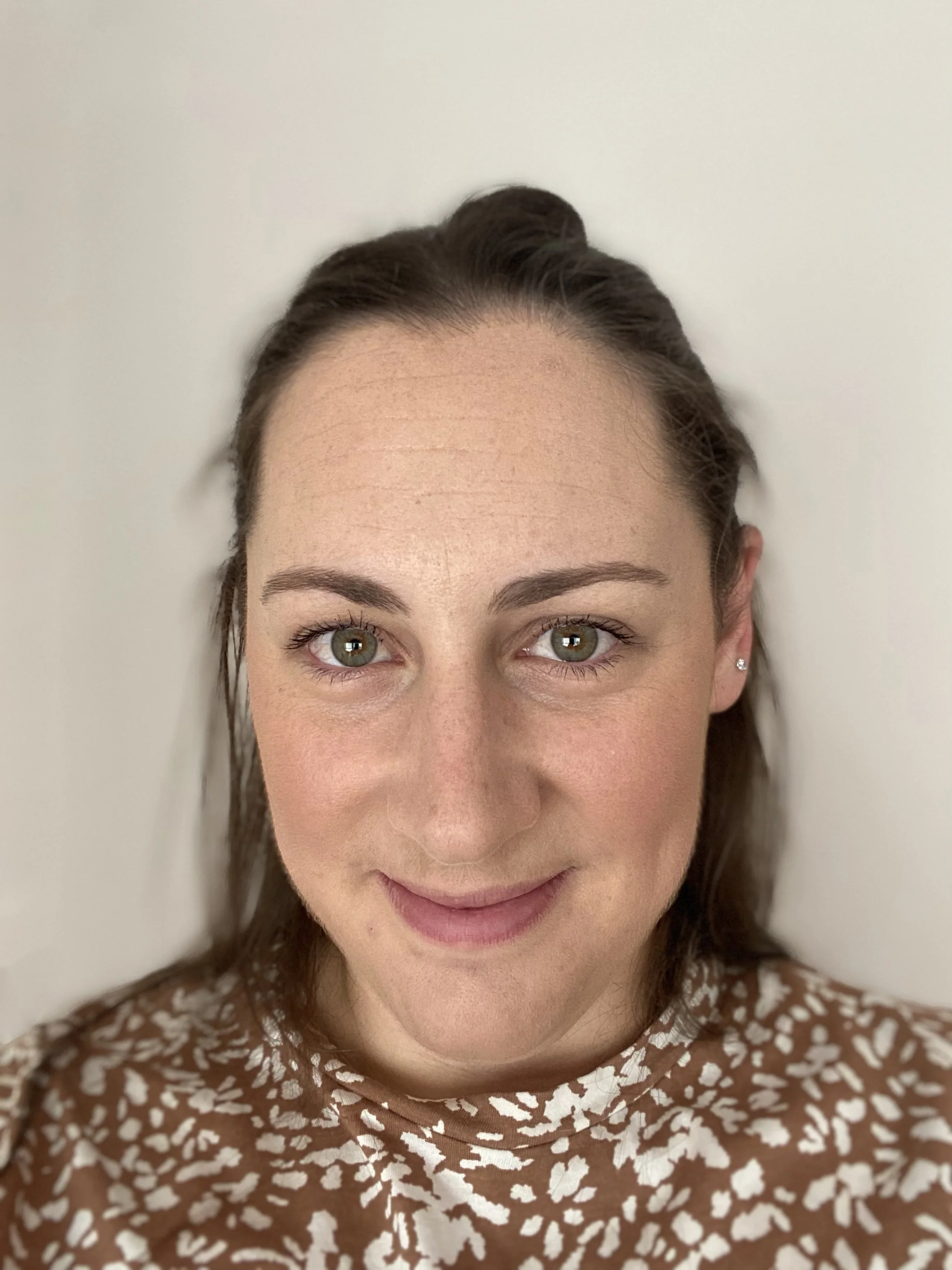 Close-up of a woman, Honnie Craig play therapist, with light skin, brown hair styled in a braid, gray-green eyes, and a warm smile, wearing a brown and white patterned top, against a plain white background.