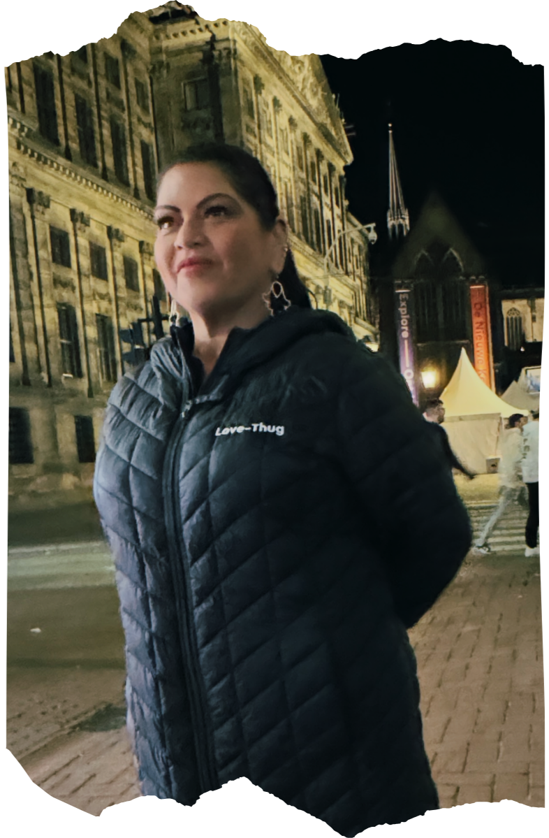 A woman standing outdoors at night in front of a historic building with ornate architecture and a spire. She is wearing a black quilted jacket with the words 'Love-Thug' on it and large earrings. There are tents and people in the background.