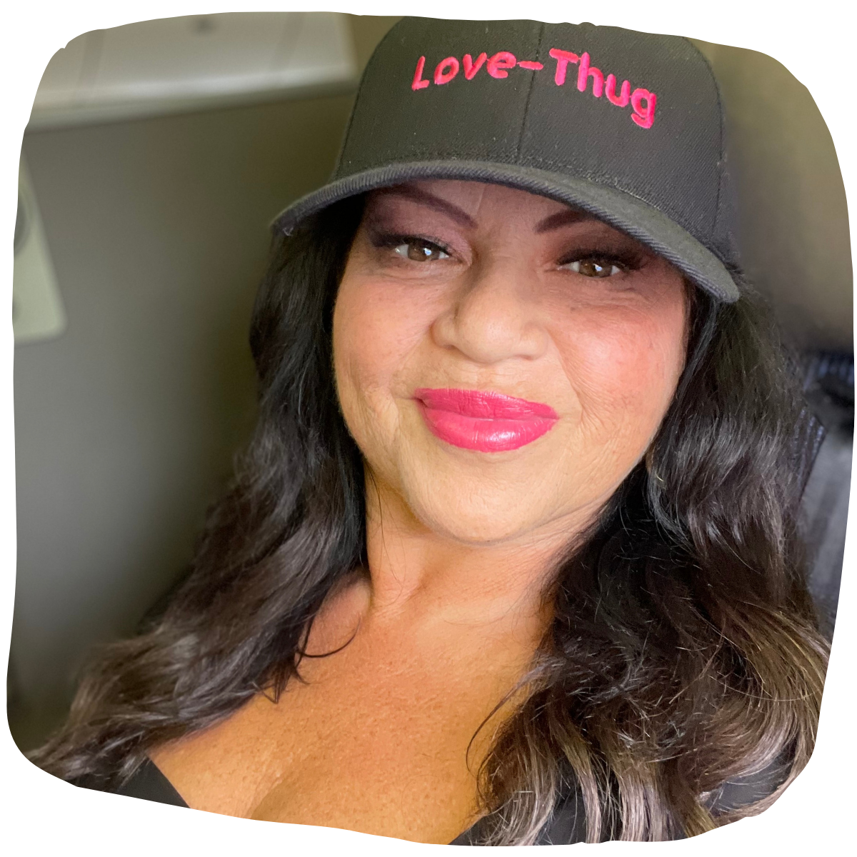 A woman with long dark hair wearing a black baseball cap with pink embroidered text that says Love-Thug, smiling with pink lipstick, inside a room.