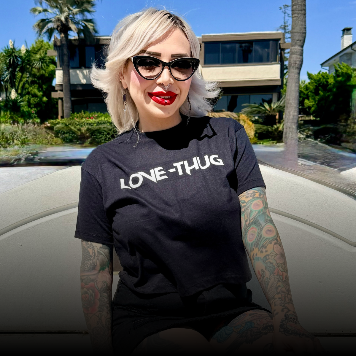 A woman with platinum blonde hair, sunglasses, bright red lipstick, and tattoos, sitting outdoors on a sunny day with a modern house and palm trees in the background. She is wearing a black T-shirt that says 'LOVE-THUG'.
