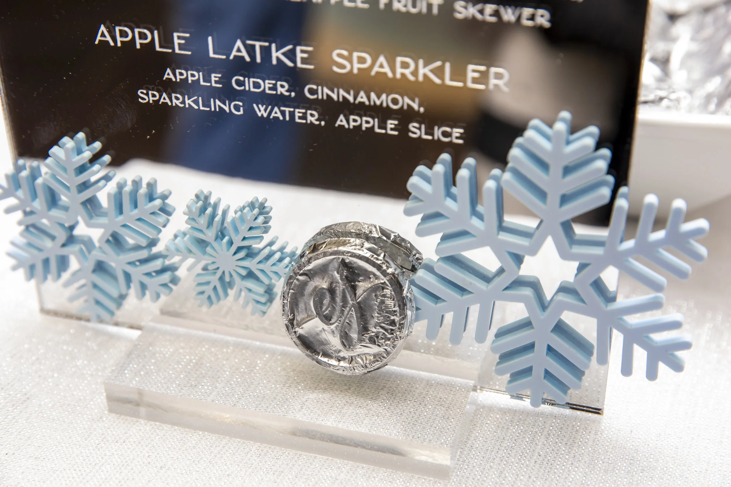 Close-up of the apple latke sparkler drink sign with blue acrylic snowflakes and a silver gelt coin