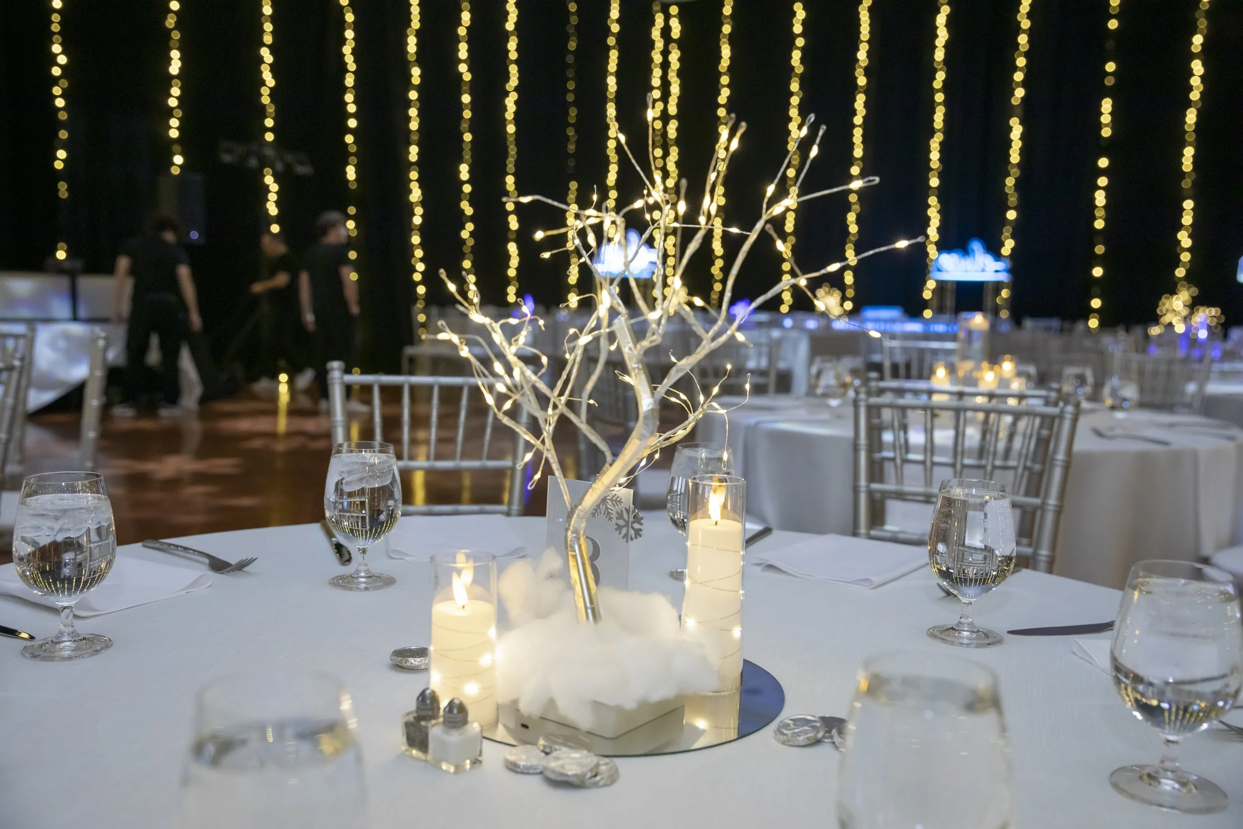 A round table centerpiece of a white branch tree with candles and faux snow, set against string lights