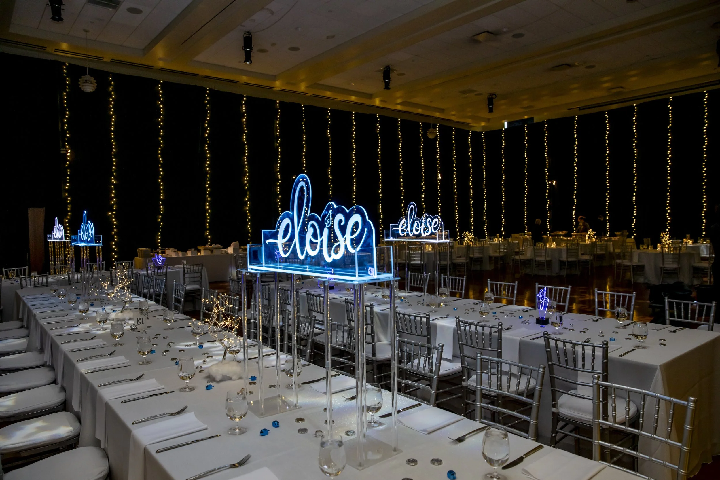 Long banquet tables set beneath a glowing eloise sign and curtains of string lights