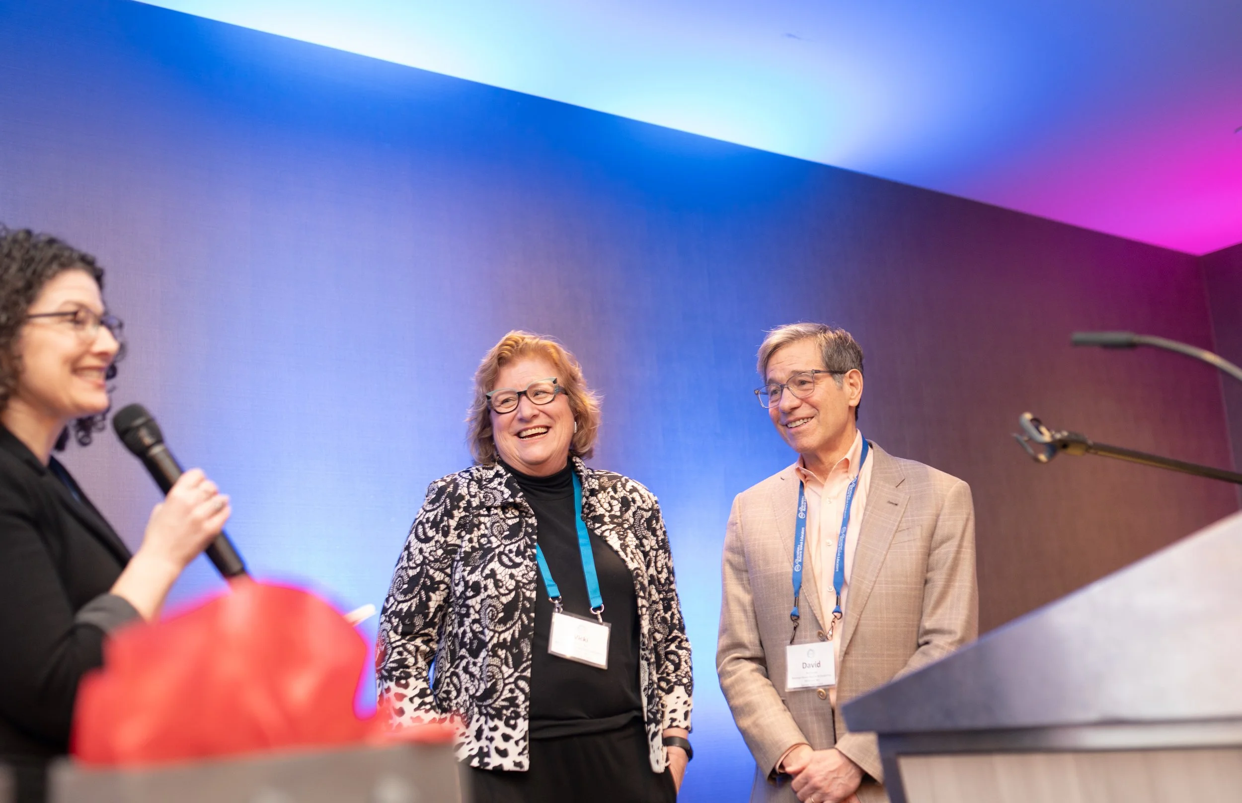 A speaker with a microphone on a blue-lit stage beside two attendees at the ARJE conference in Minneapolis