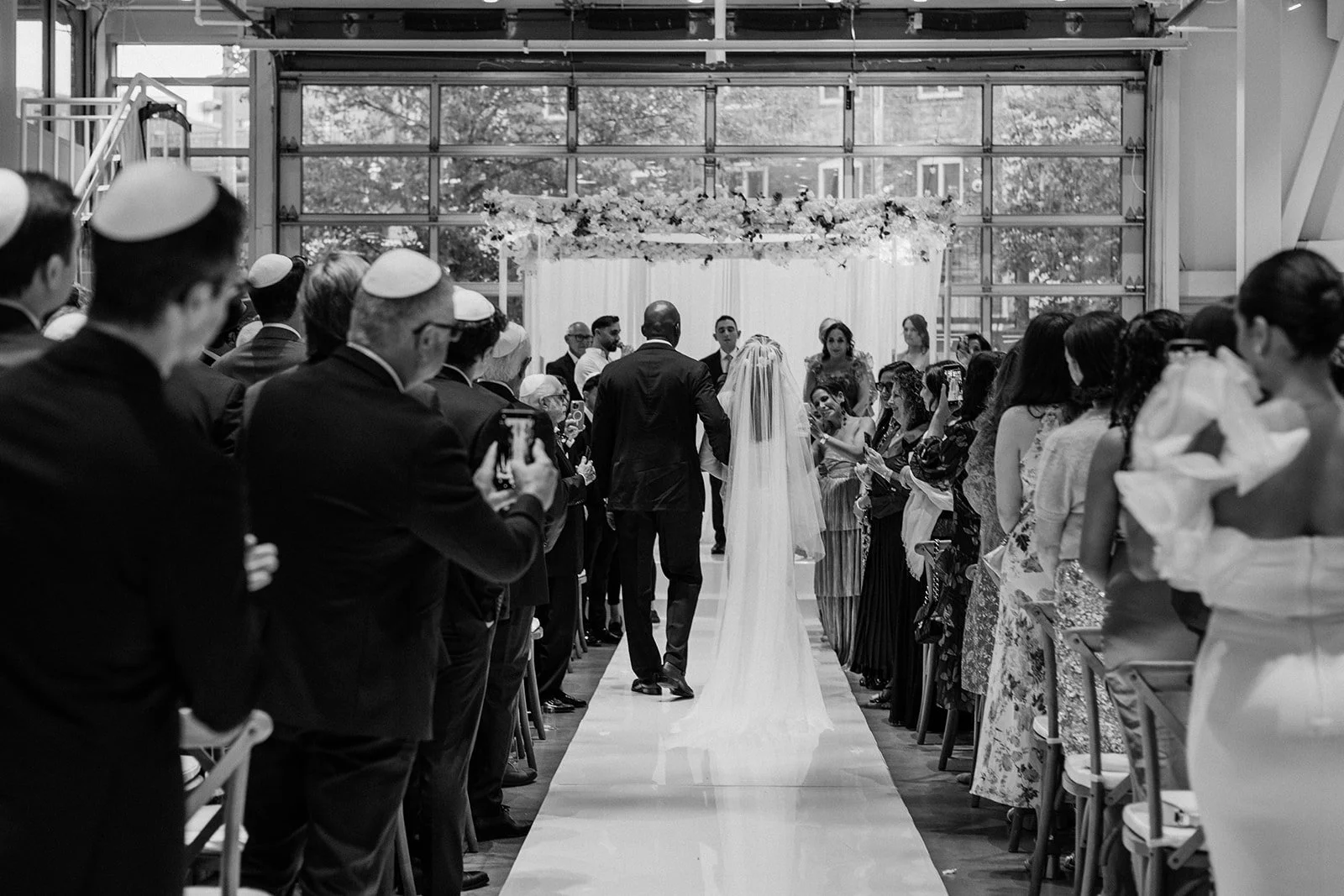 Black and white photo of the bride walking down the aisle between standing guests toward the chuppah
