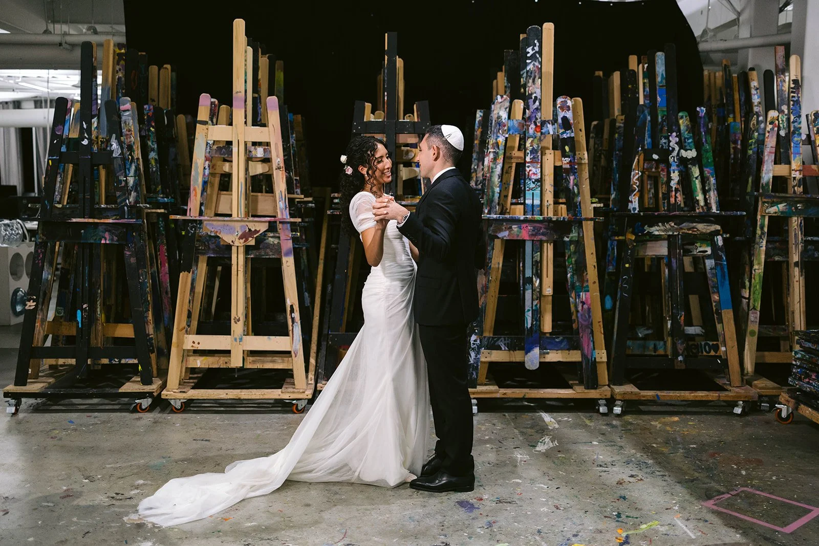 The couple embraces among rows of wooden artists' easels in the gallery studio