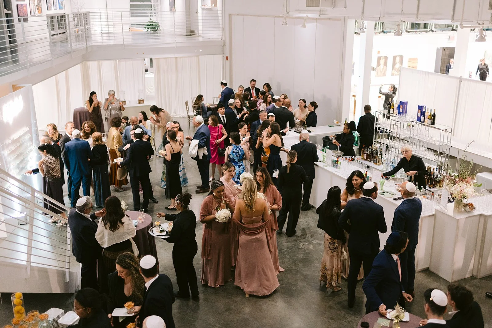 Guests and bridesmaids in dusty rose mingle at the bar in the bright white gallery during cocktail hour